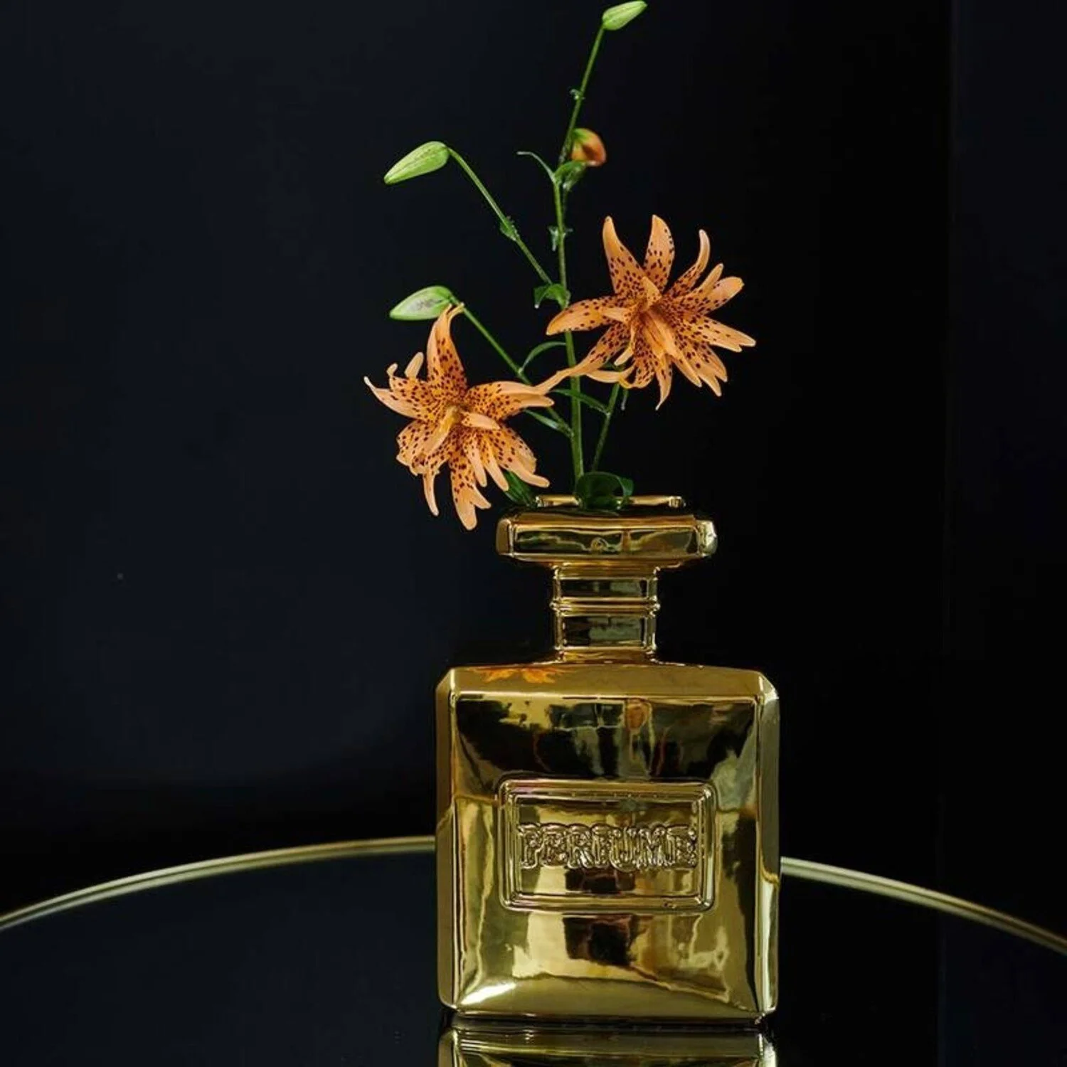 Pink and orange tiger lily flowers in a gold-colored perfume bottle against a black background.
