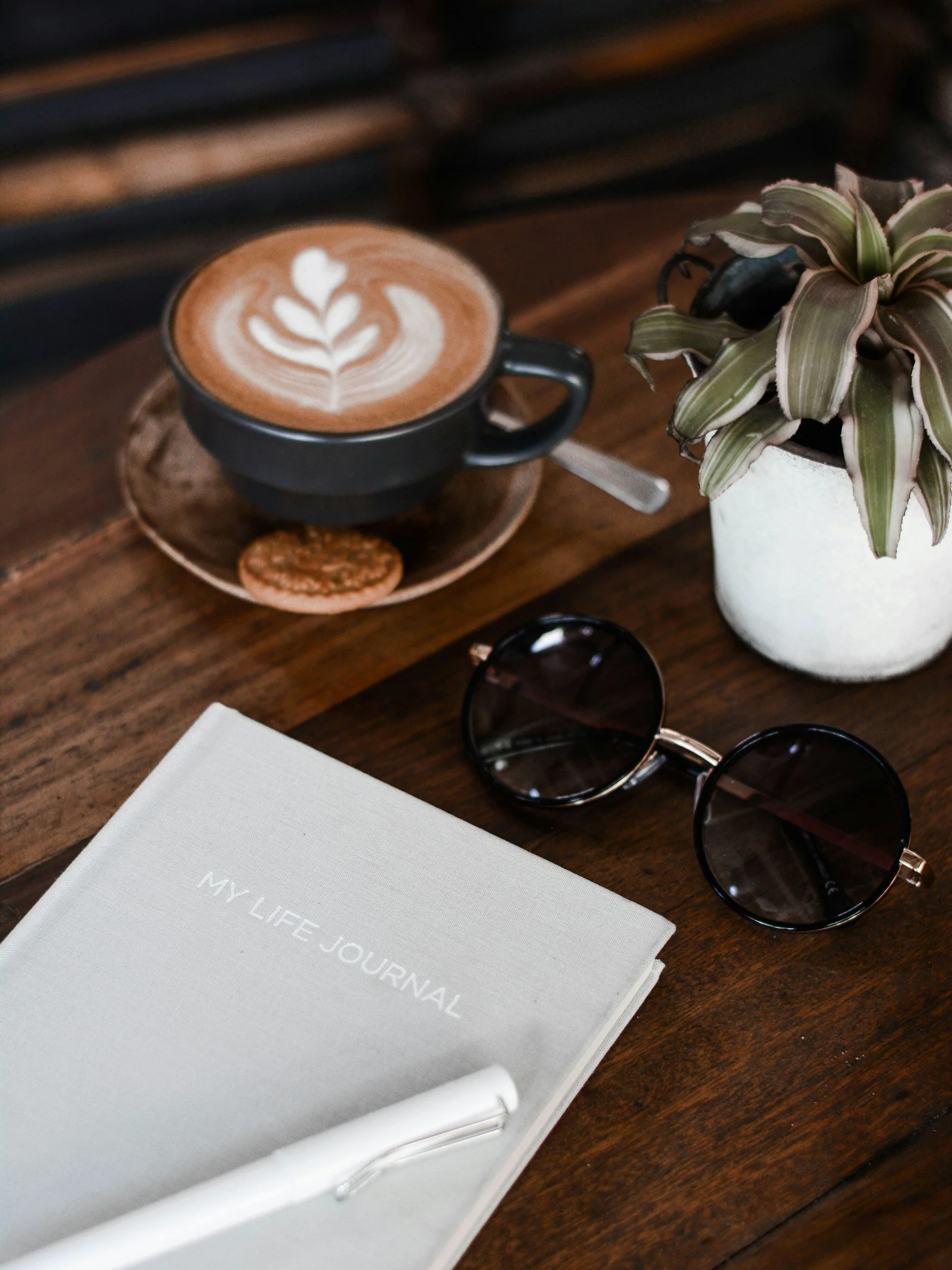 A white journal titled 'My Life Journal' on a wooden table, next to a pair of sunglasses, a latte with latte art, a cookie on a saucer, and a potted plant.