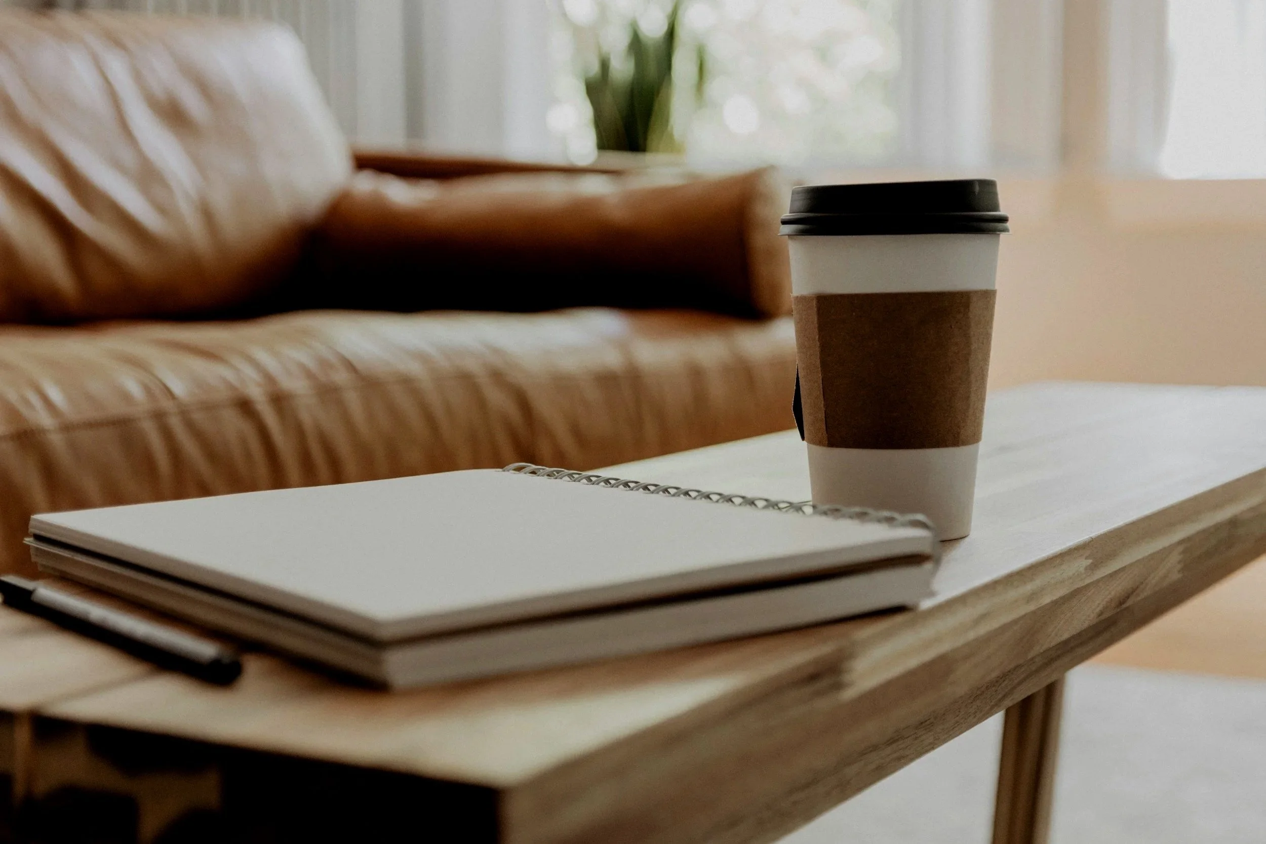 A disposable coffee cup with a brown paper sleeve and black plastic lid on a wooden table, with a closed notebook and a pen in the foreground, and a blurred background showing a window and green plant.