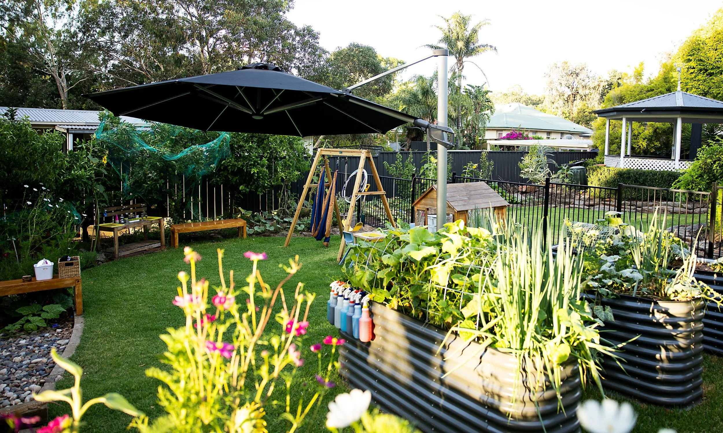 A backyard garden with a lawn, flower beds, a large dark patio umbrella, a wooden swing set, and a small wooden playhouse, surrounded by trees and a black fence.