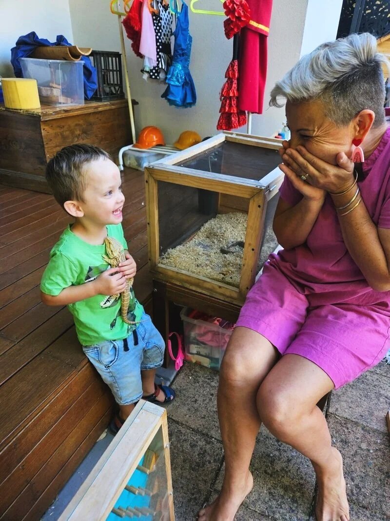 A young boy holding a lizard and smiling at an older woman with short, gray hair who is laughing and covering her mouth with her hands. They are indoors with wooden and brick flooring, beside a terrarium with sand and a small animal, likely a reptile.