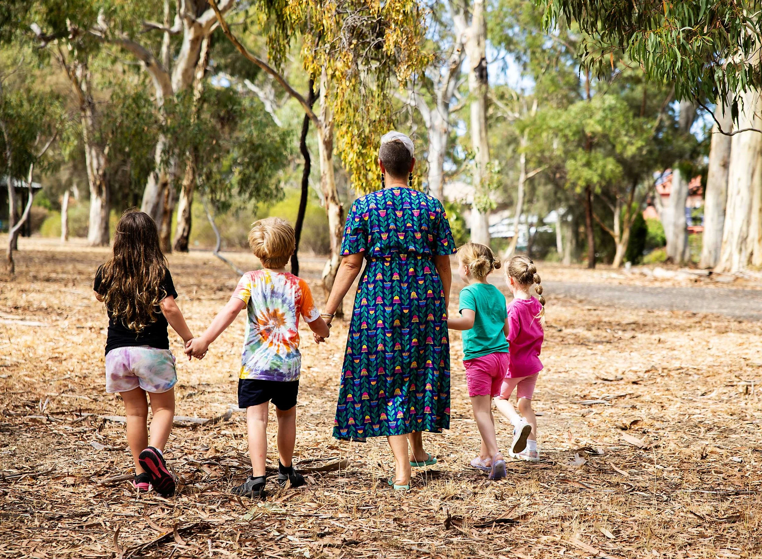 A woman with grey hair and earrings dressed in a colorful patterned dress is walking hand-in-hand with a group of children in a wooded park. The children wear casual clothes, some with tie-dye shirts, and are walking on a dirt path covered with falle