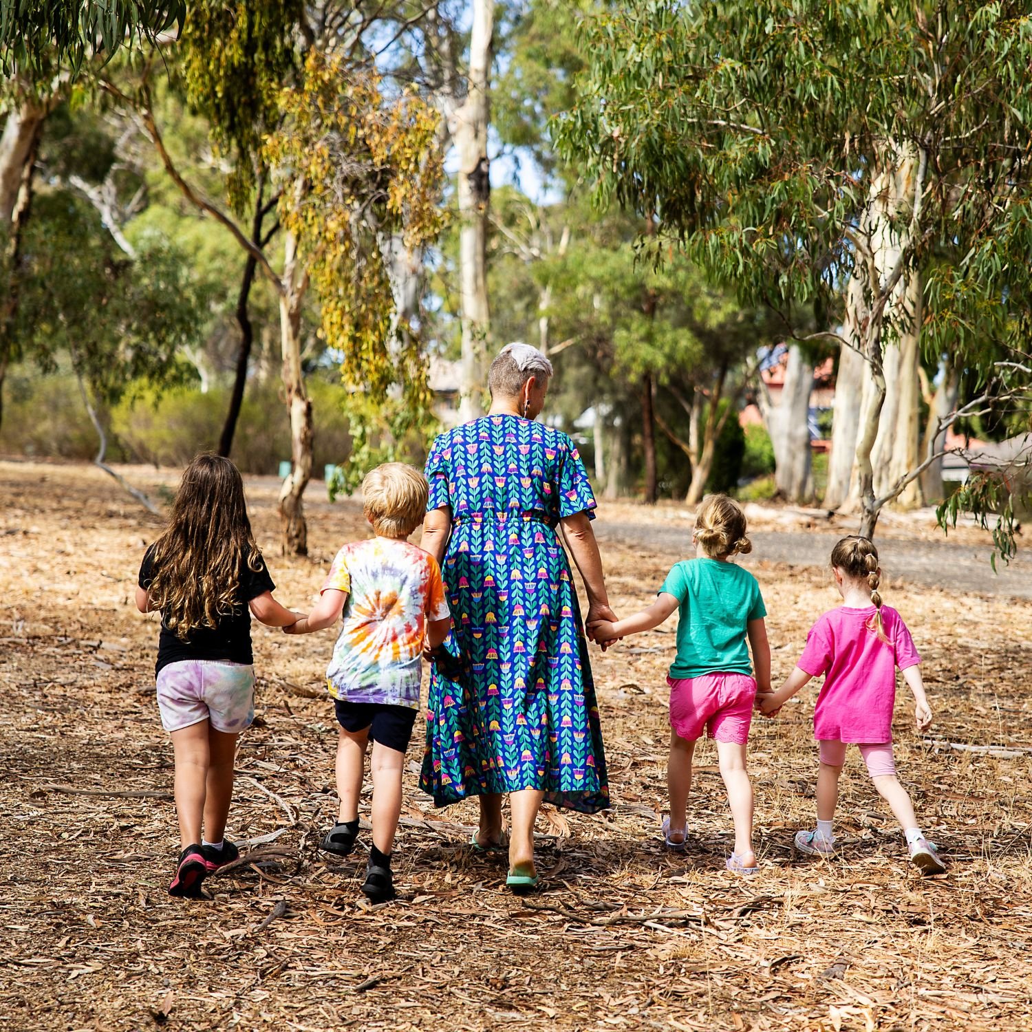 A woman walking hand-in-hand with five children through a wooded area, surrounded by trees and dry leaves on the ground.