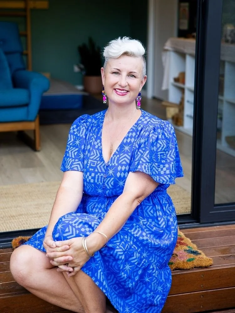 A woman with short, platinum blonde hair sitting on a wooden porch in front of sliding glass doors. She is wearing a blue patterned dress and pink earrings, smiling at the camera.