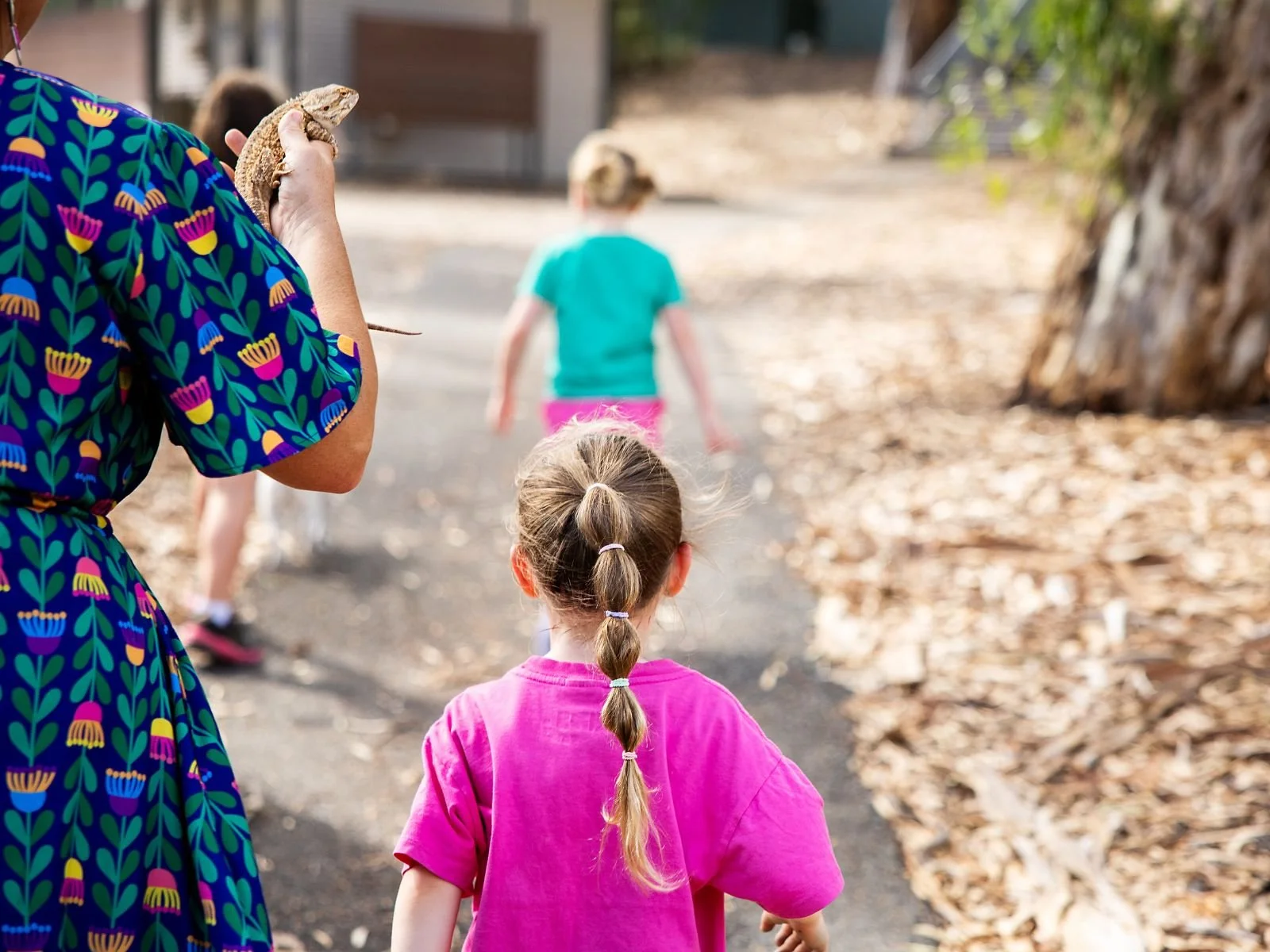 A woman holding a bearded dragon lizard on her hand while walking with children outdoors on a dirt pathway lined with fallen leaves, with trees and blurred structures in the background.