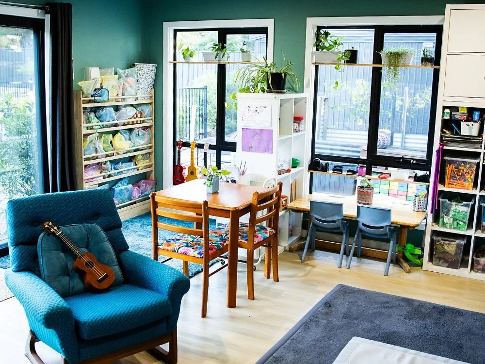 A cozy room with a teal wall, featuring a wooden dining table with four chairs, a blue armchair with a ukulele on it, and a white bookshelf. Large windows with plants on the sill allow natural light, and there are storage shelves with books and toys.