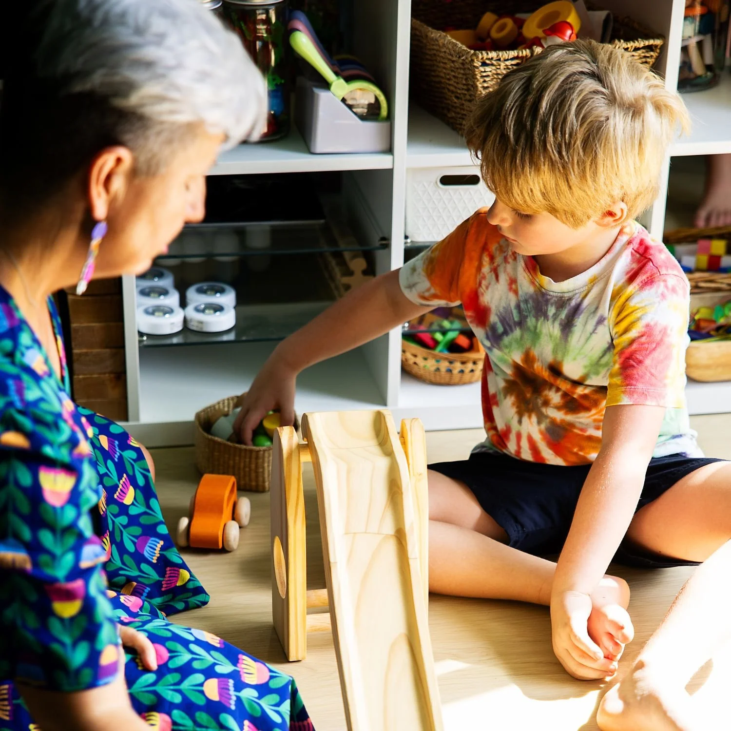 A young boy and a woman sitting on the floor playing with wooden toy blocks in a playroom. The woman is wearing a colorful dress with a floral pattern, and the boy is wearing a tie-dye T-shirt and black shorts. Shelves with toys and books can be seen in the background.