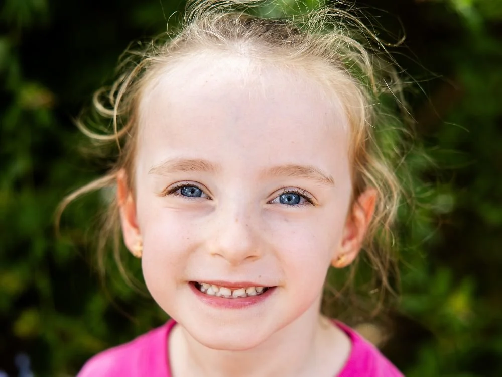 Close-up of a smiling young girl with blonde hair and blue eyes, wearing a pink shirt, outdoors with green foliage in the background.