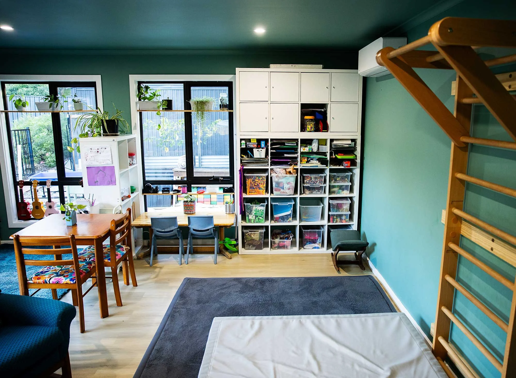 A cozy living room with large windows, white storage shelves filled with toys and books, a wooden dining table with colorful chairs, a dark blue armchair, and a small black step stool, all on wooden flooring with a grey rug.