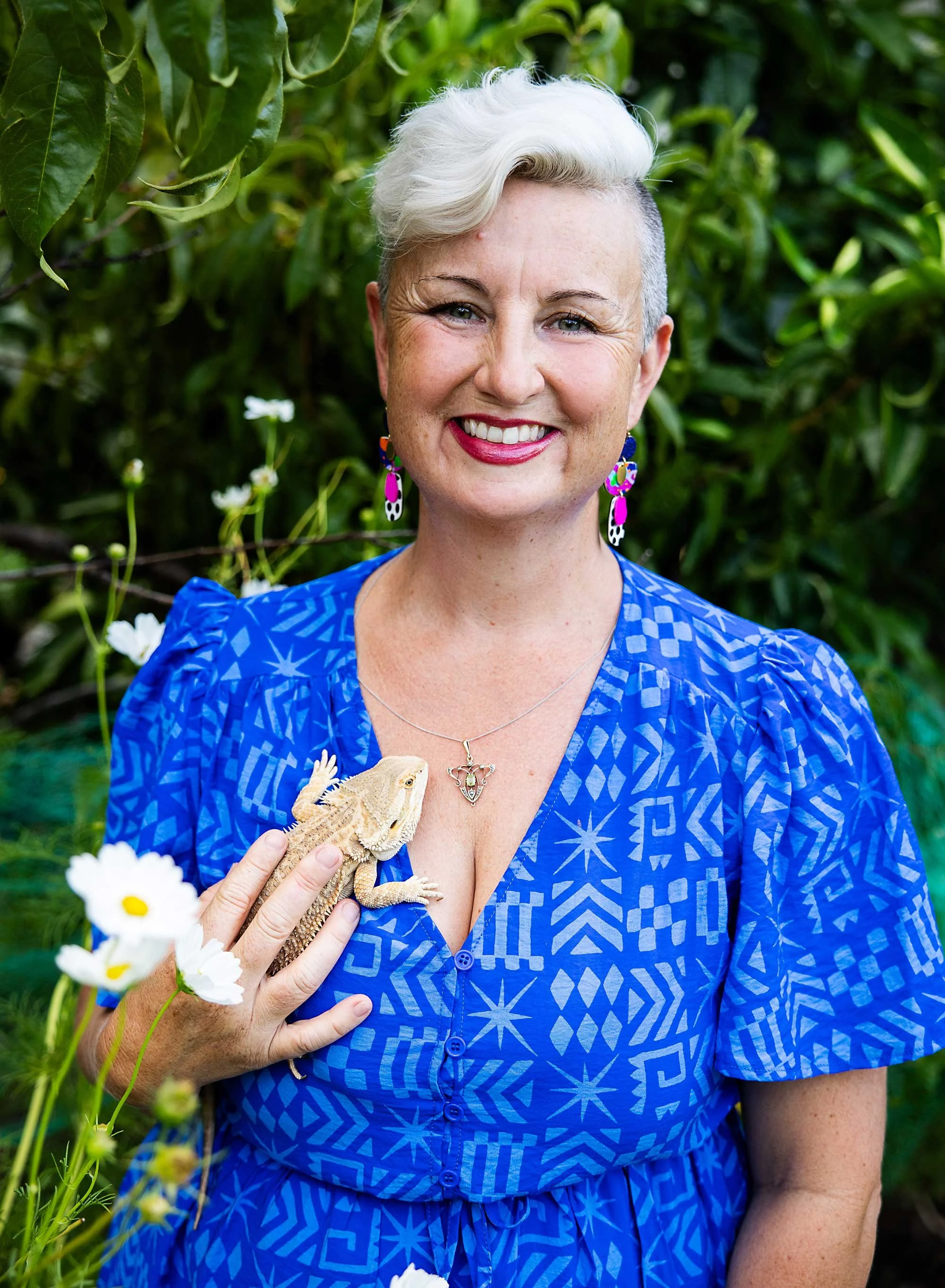 A woman with short, platinum blonde hair and fair skin smiling outdoors, holding a bearded dragon lizard on her hand, surrounded by green foliage and white flowers.