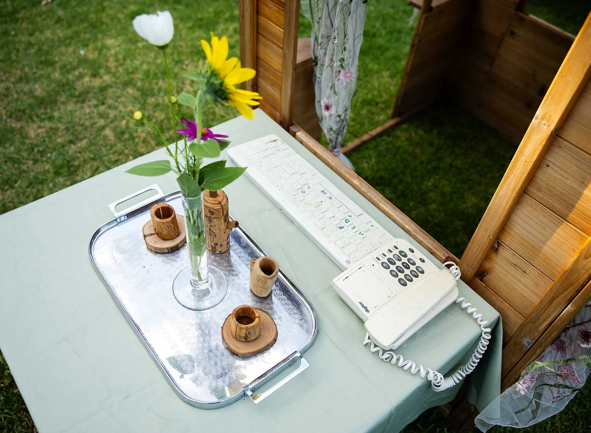 A table outdoors with a vase of flowers, a tray with wooden candle holders, a white keyboard, and a white landline phone.