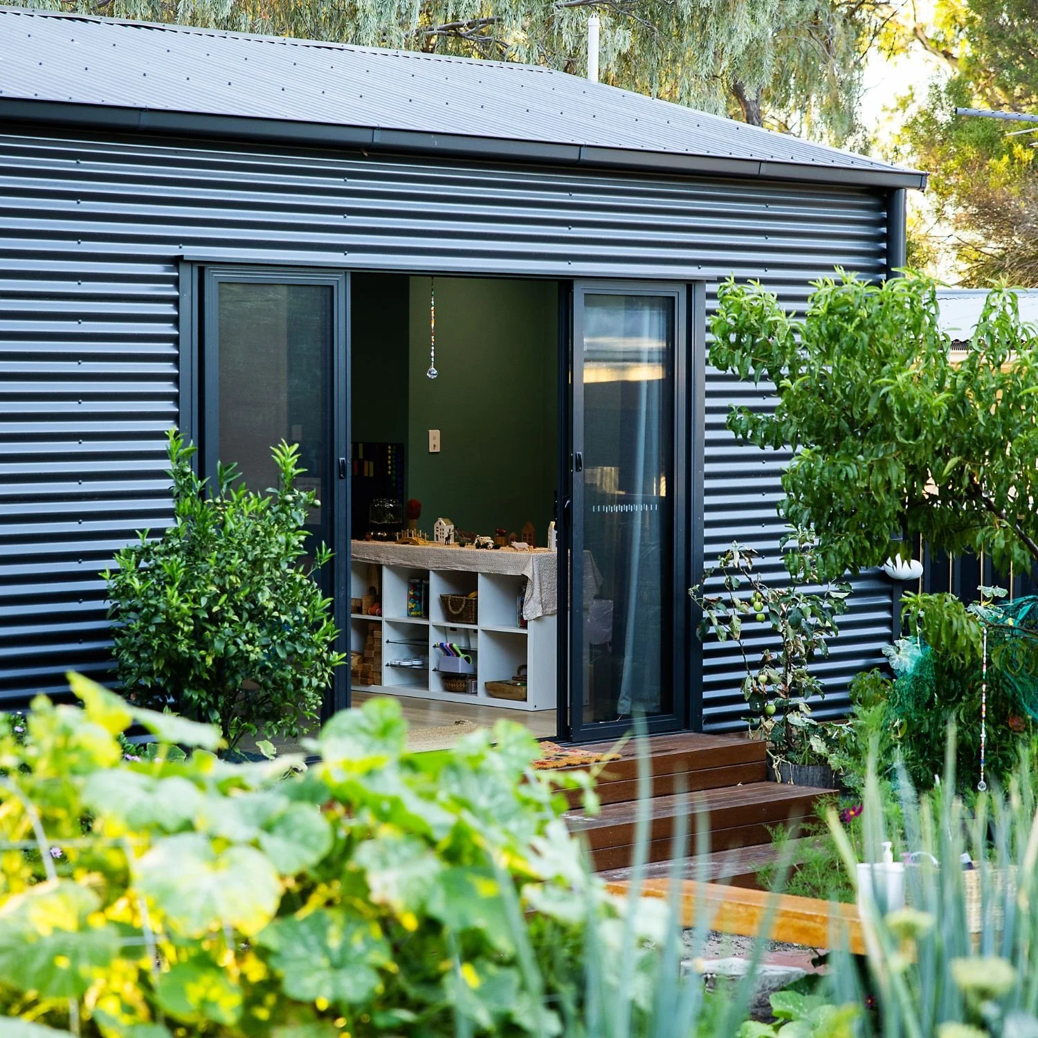 A house with black metal siding has an open sliding glass door leading to a room with green interior walls. Inside, there is a white table with toys and decorations, and open cubbies underneath. The house is surrounded by greenery and plants, with a weathered wooden deck and a garden area in the foreground.
