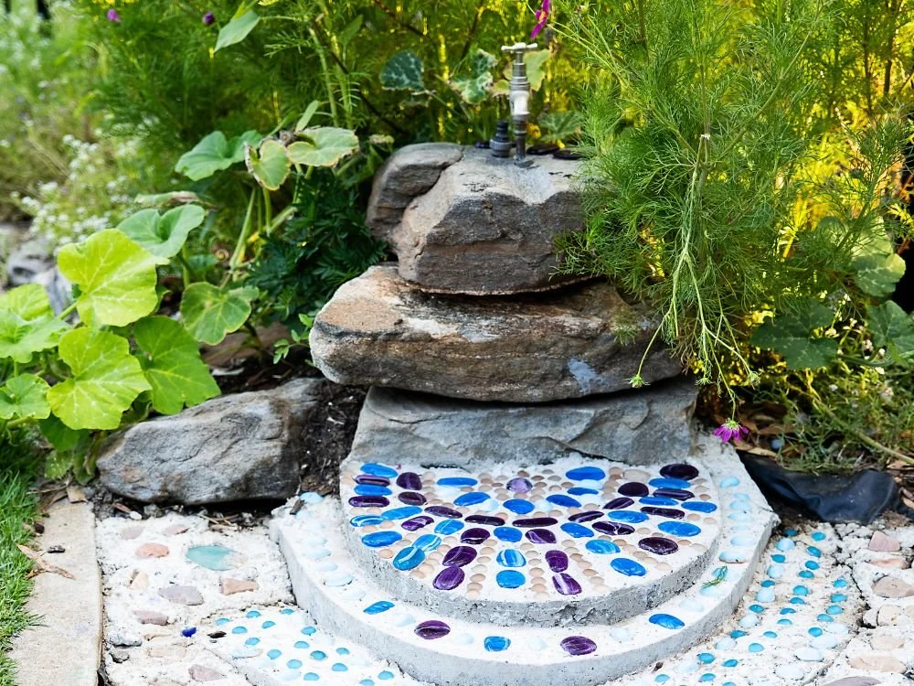 Garden fountain with stacked rocks, mosaic pebble designs, surrounded by green plants and flowers.