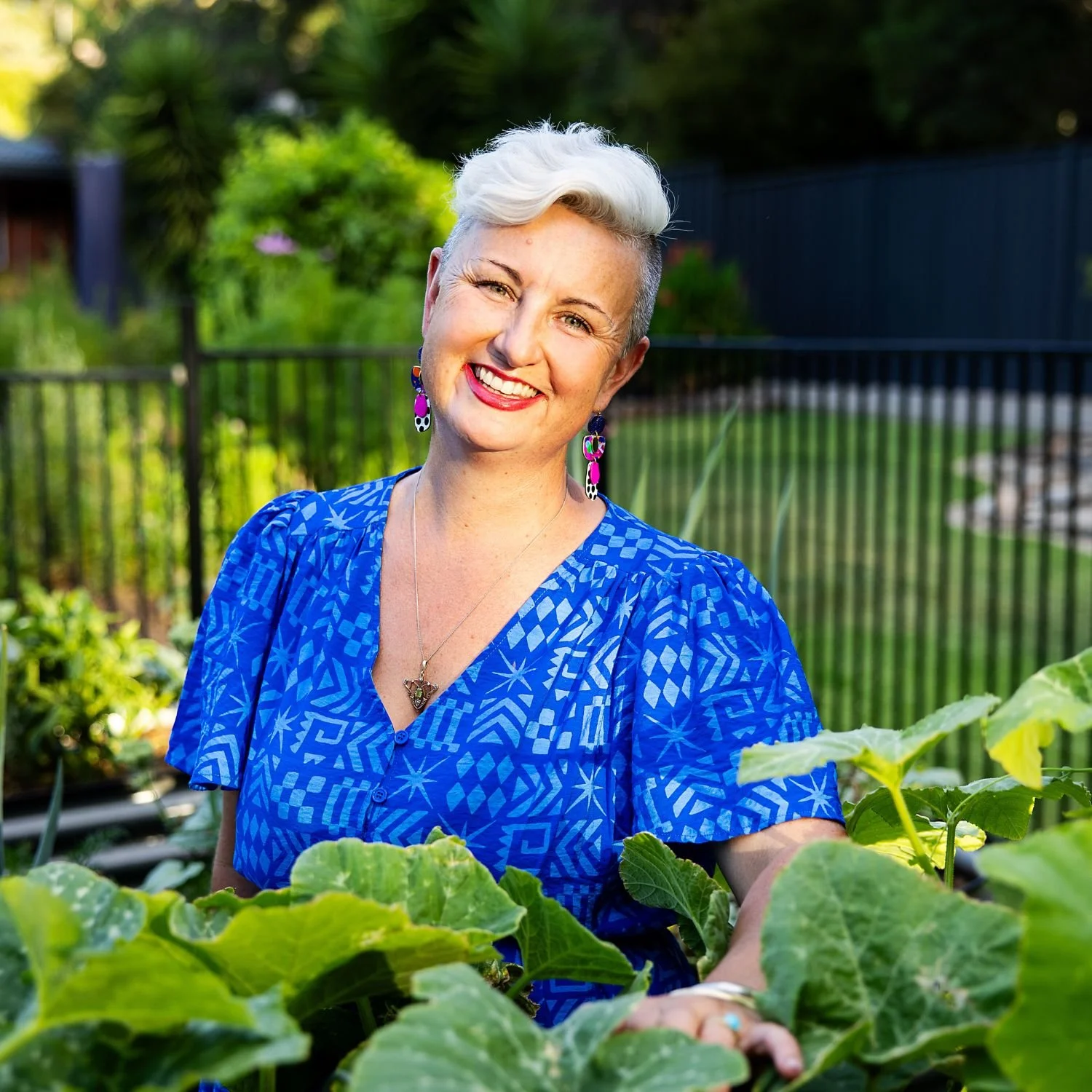 A smiling woman with short white hair wearing a blue patterned shirt, colorful earrings, and a necklace, standing in a garden among green plants and sunlight.