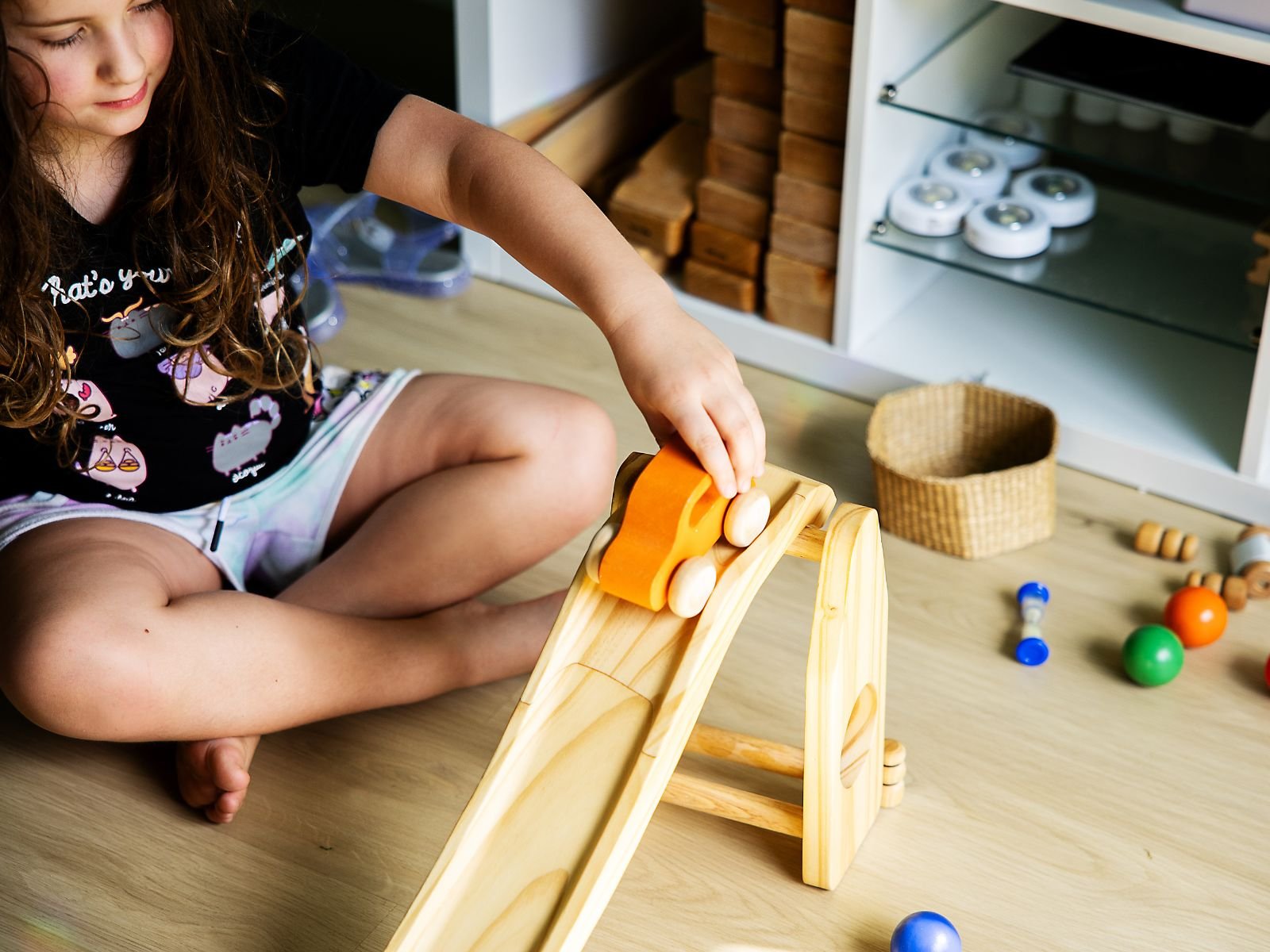 A young girl sitting cross-legged on a wooden floor, playing with a wooden toy car ramp and orange and white cars, with colorful marbles and a small basket nearby.