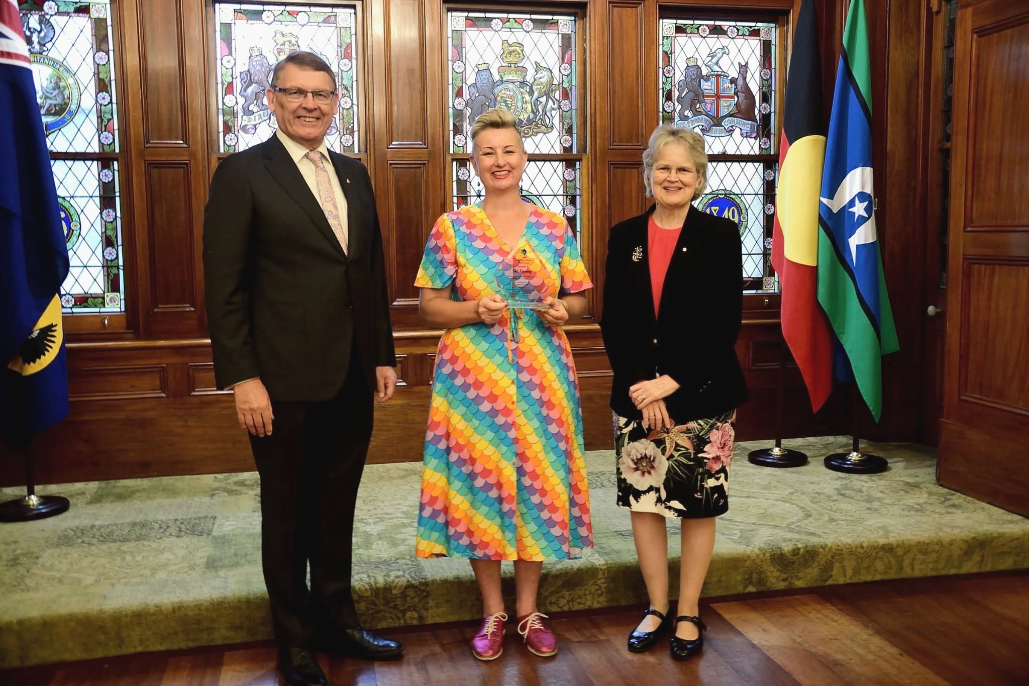 Three people standing indoors in front of stained glass windows and flags. The man on the left is wearing a black suit, white shirt, and multicolored tie. The woman in the middle is wearing a colorful, patterned dress and holding an award. The woman on the right is wearing a black jacket and floral skirt.