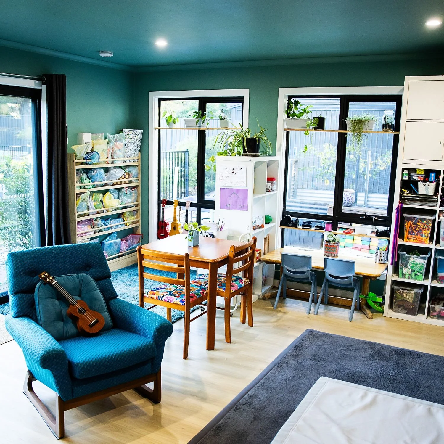 Interior of a family room with green walls, large windows, a blue armchair with a ukulele on it, a wooden dining table with colorful chairs, shelves with plants and toys, and a play area.