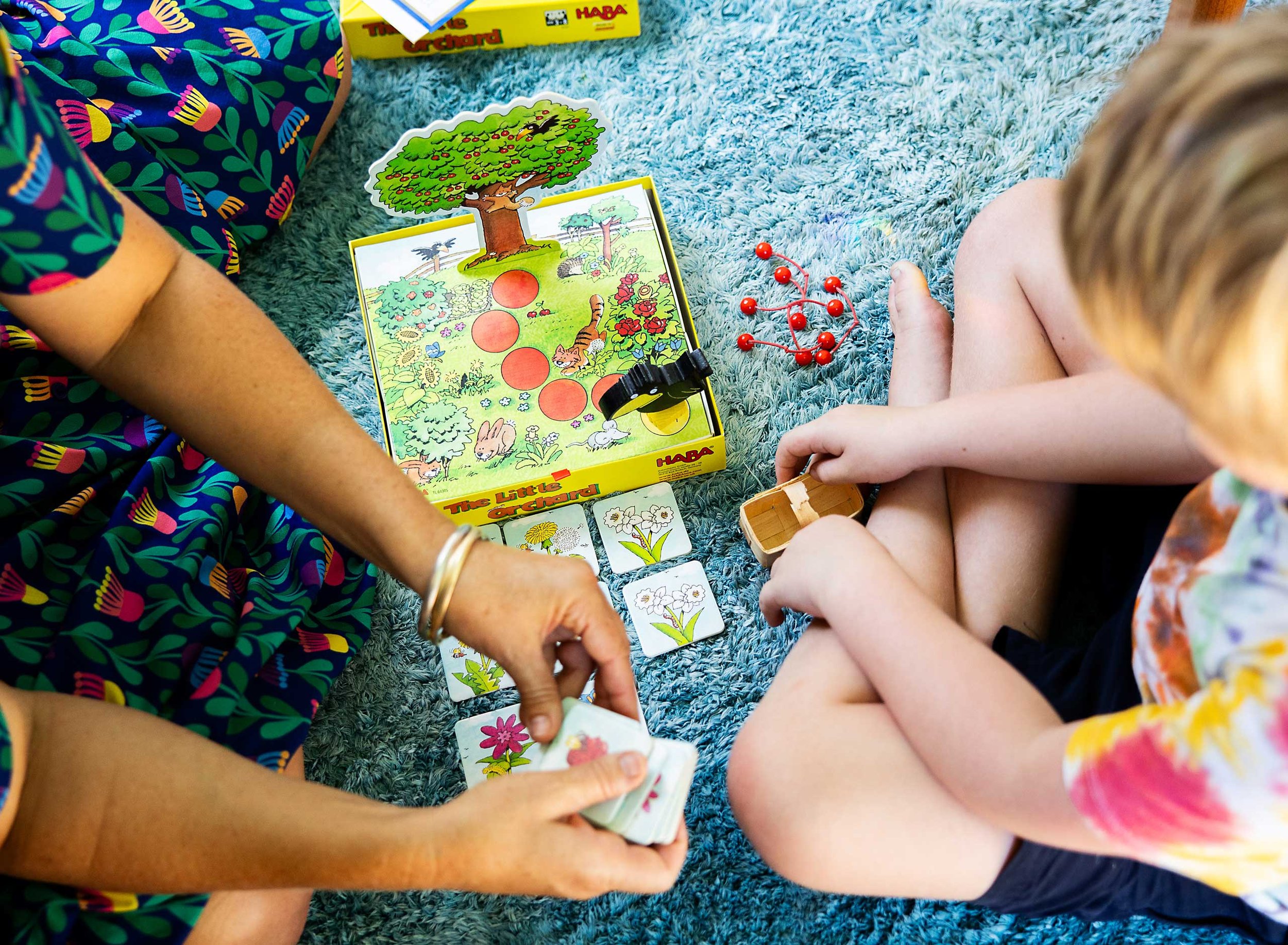 Two children playing a card game on a blue rug, with a board game box partially visible, and various illustrated cards with flowers and animals.