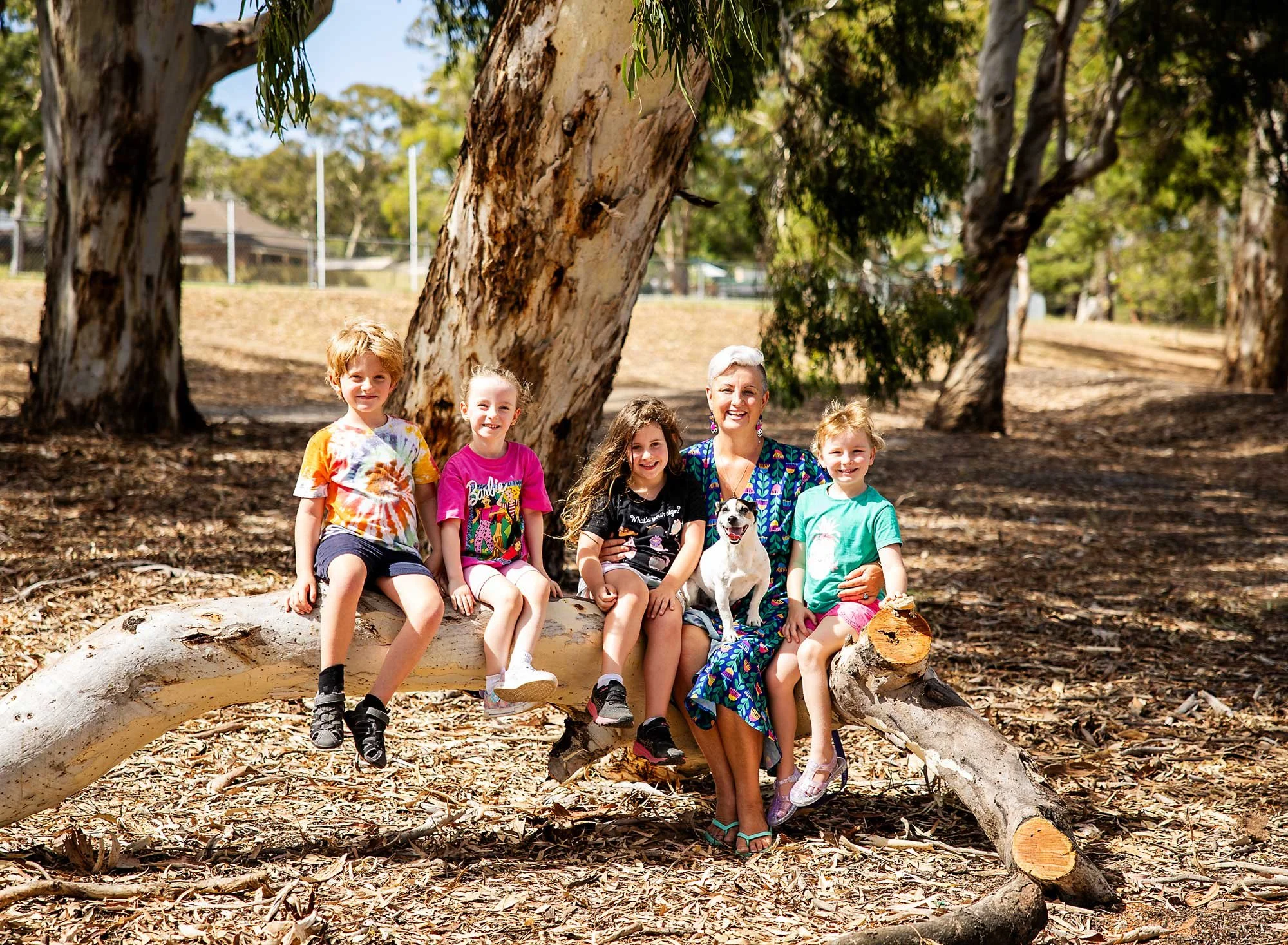 Six children and one woman sitting on a large fallen tree trunk in a park, with trees and a fence in the background.