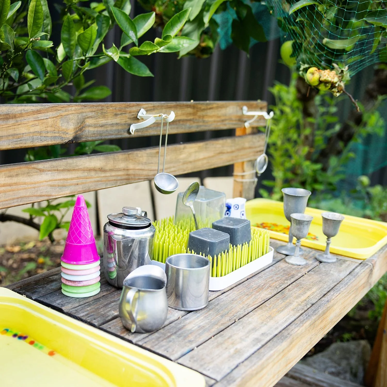 Outdoor party table with colorful cups, metal cups, drink dispenser, and drinking accessories on a wooden table with a wooden privacy fence and green foliage in the background.