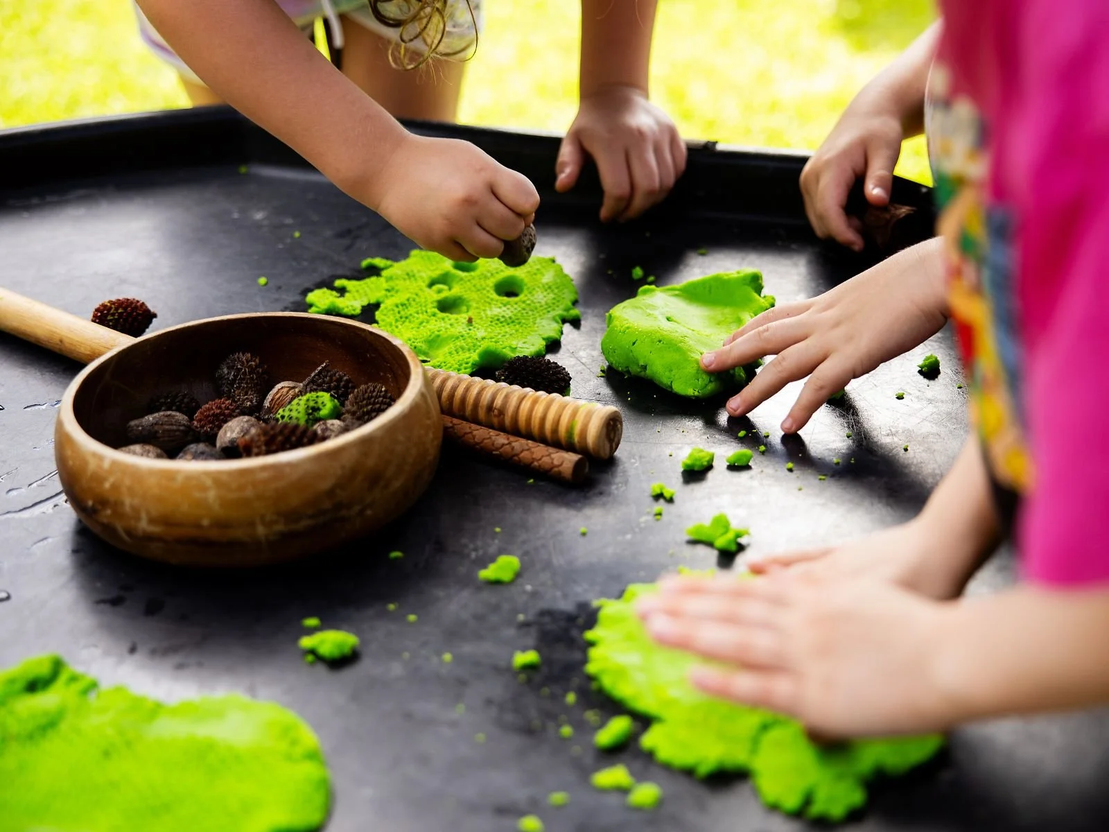 Children playing with bright green playdough on a black table outdoors, with scattered pieces and a wooden bowl filled with pinecones nearby.