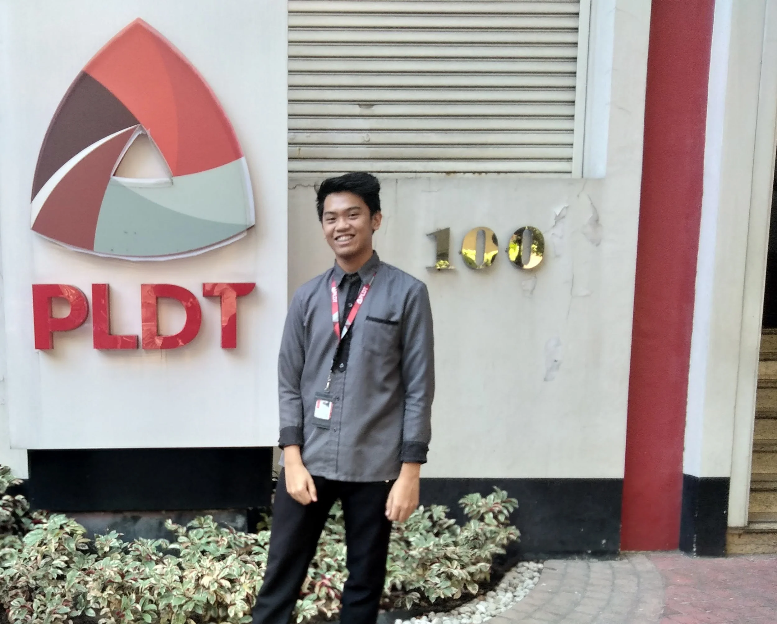 A smiling young man in a gray shirt and black pants standing outdoors in front of a sign that reads 'PIDT' and a reflective '100' sign on a textured wall with plants at the bottom.