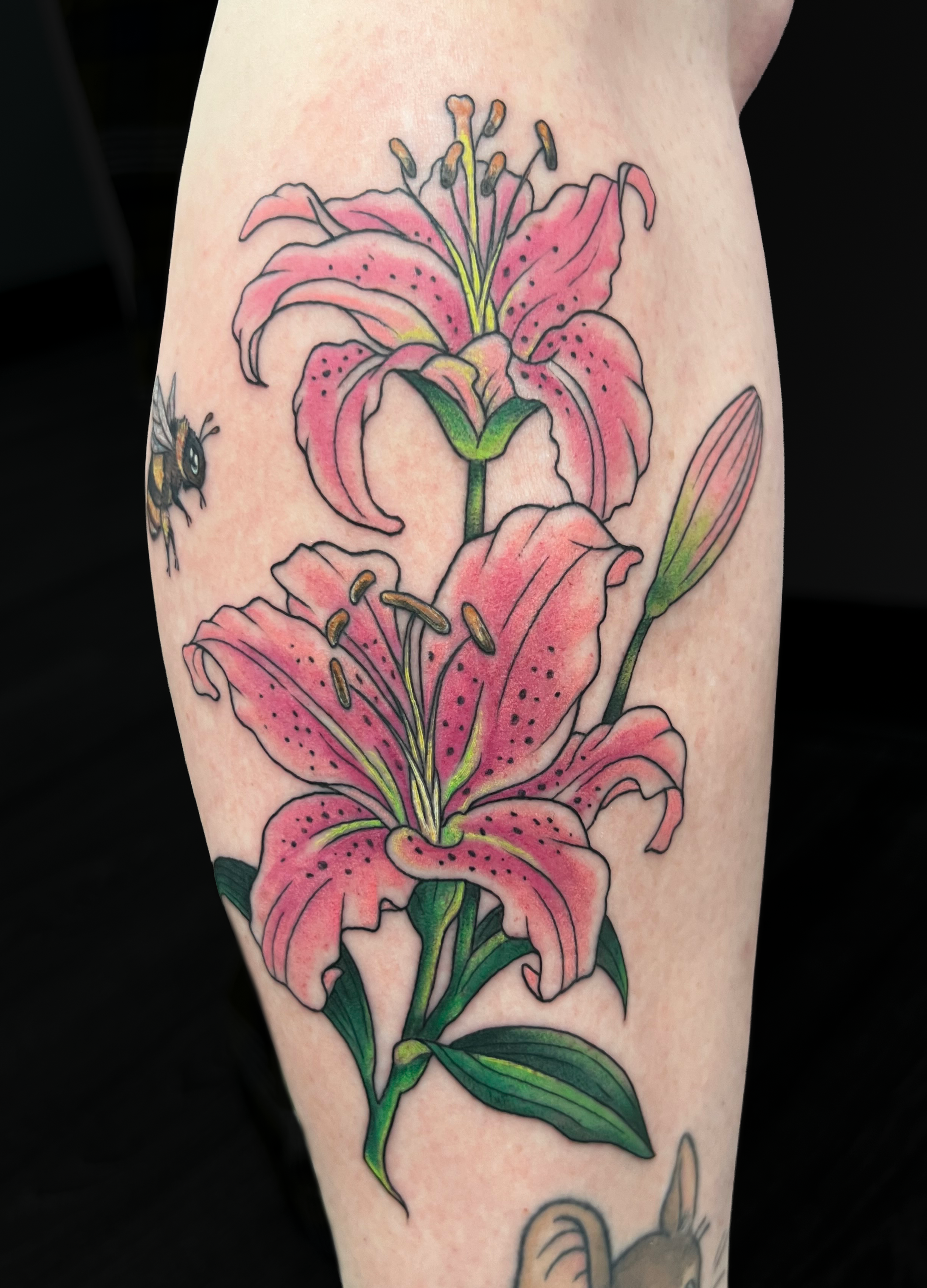 A tattoo of a pink lily flower with green leaves on someone’s skin, with a bee near the flower.