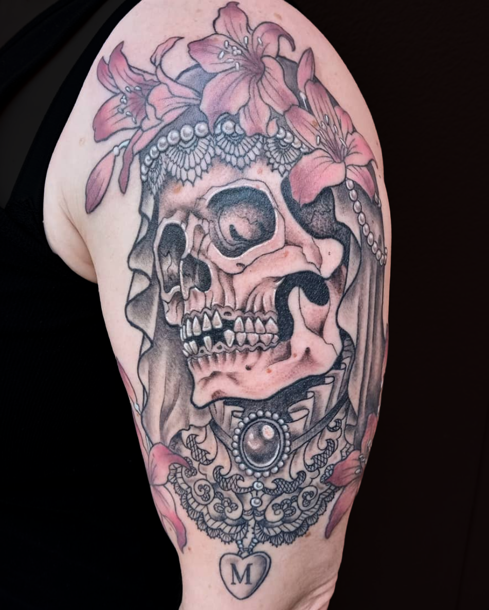Tattoo of a skull adorned with pink flowers, pearls, and a necklace featuring a heart with the letter 'M' on it.