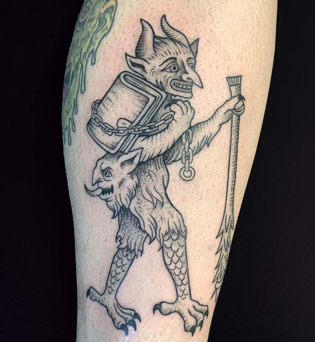 Line art tattoo of a hybrid creature with a human-like body, a goat's head, wings, and a lion's head on its chest, holding a stick in one hand and a chain in the other.