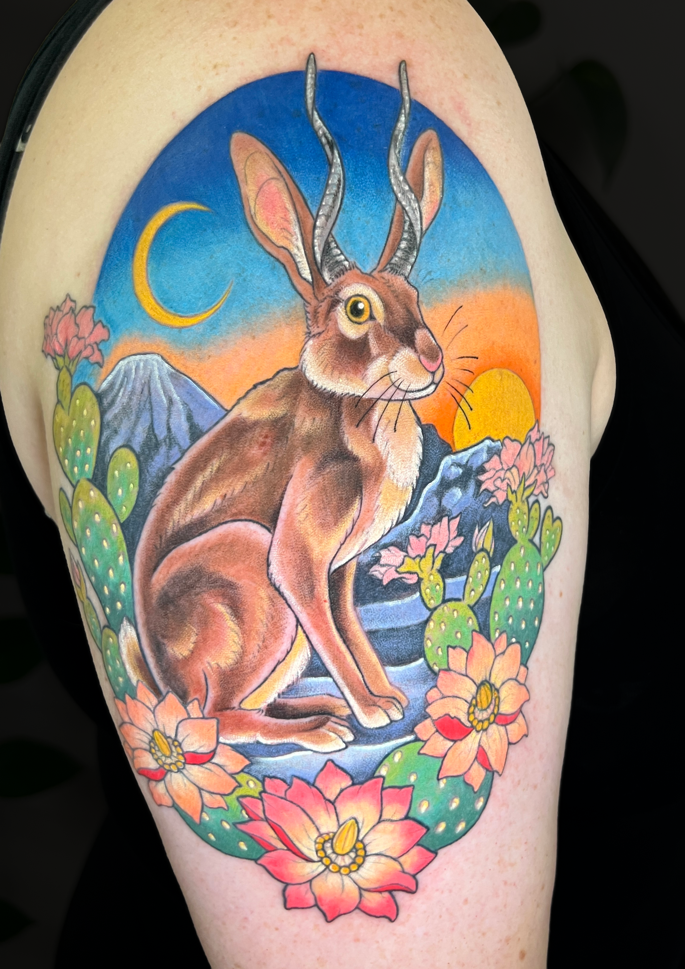 Colorful tattoo of a jackrabbit with large ears and curved antelope horns, surrounded by desert flowers, cacti, mountains, a yellow crescent moon, and a setting sun on upper arm.