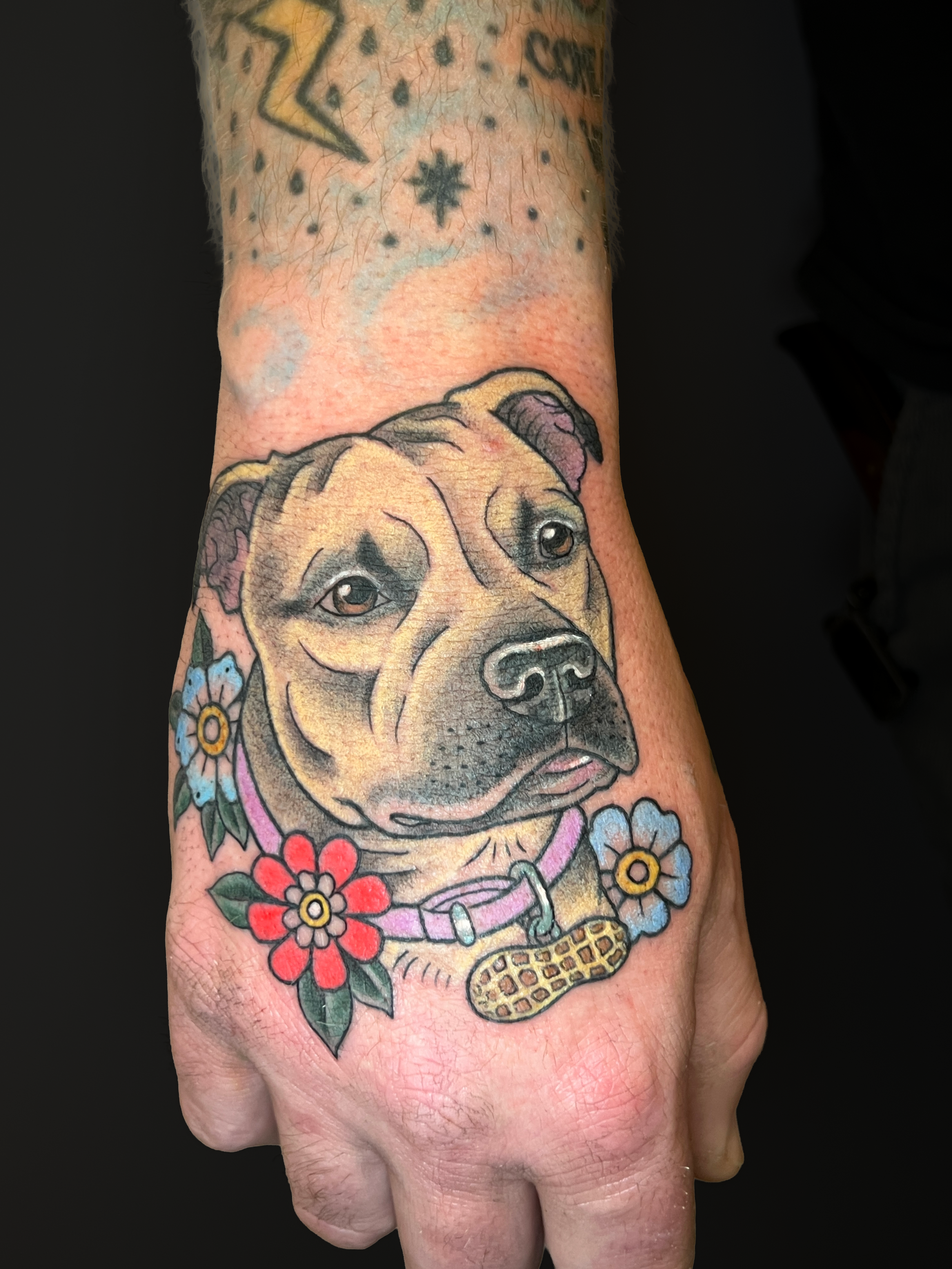 Colorful tattoo of a dog's face surrounded by flowers on a person's hand and wrist.