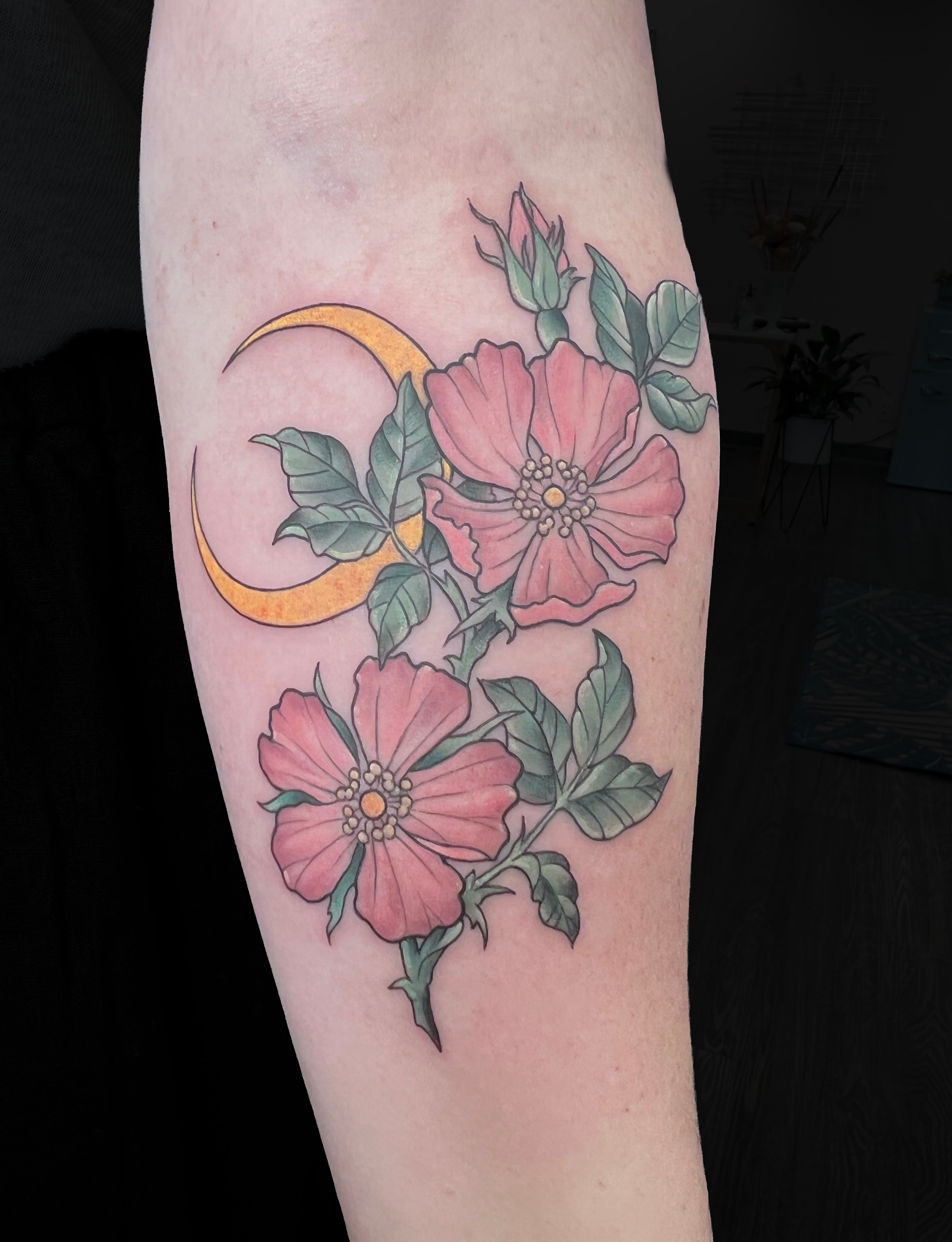 Colorful tattoo of pink flowers, green leaves, and a yellow crescent moon on a person's arm.