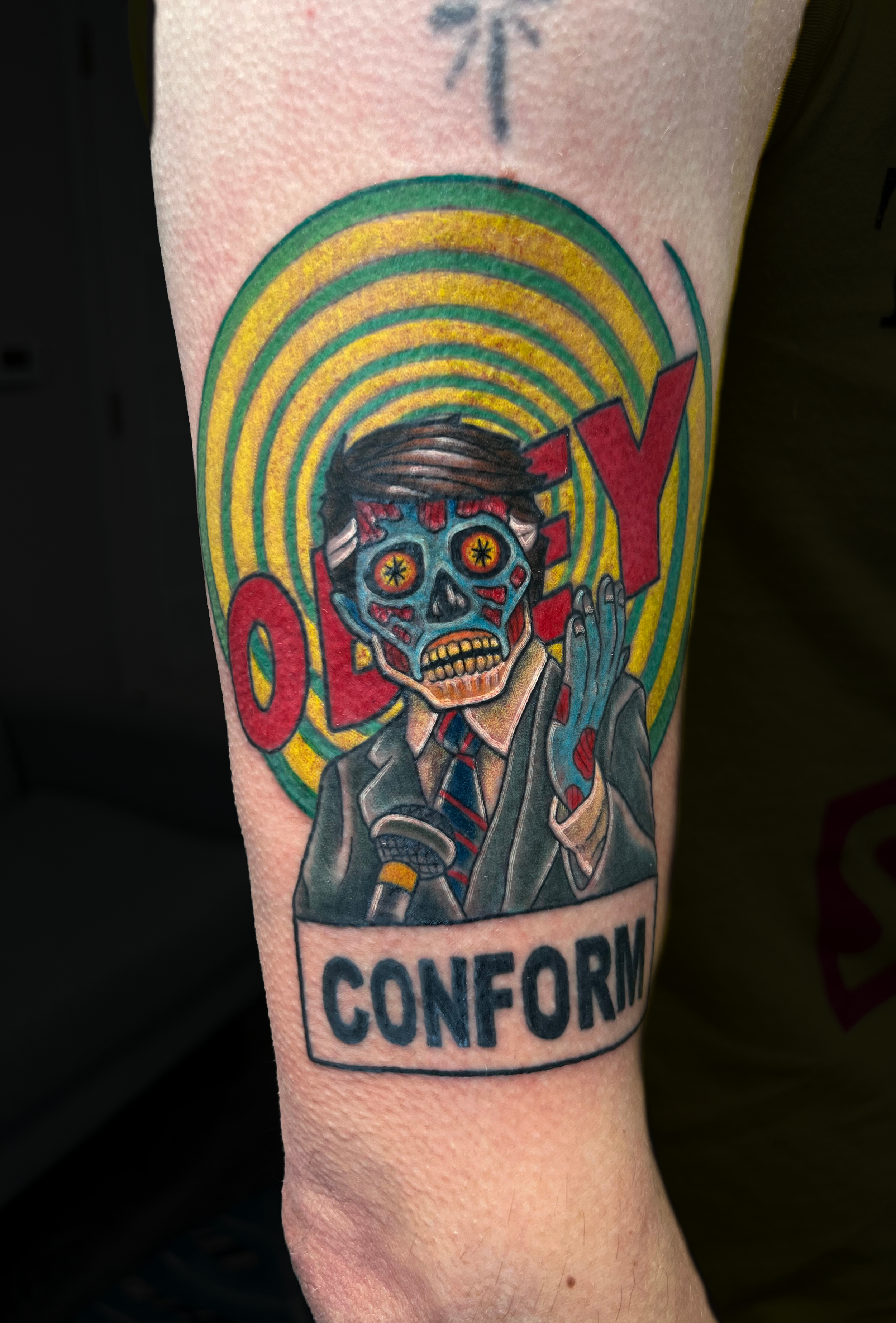 Tattoo of a skeleton in a suit with a tie, holding a marijuana joint, with rainbow-colored concentric circles in the background, and the word "CONFORM" at the bottom.