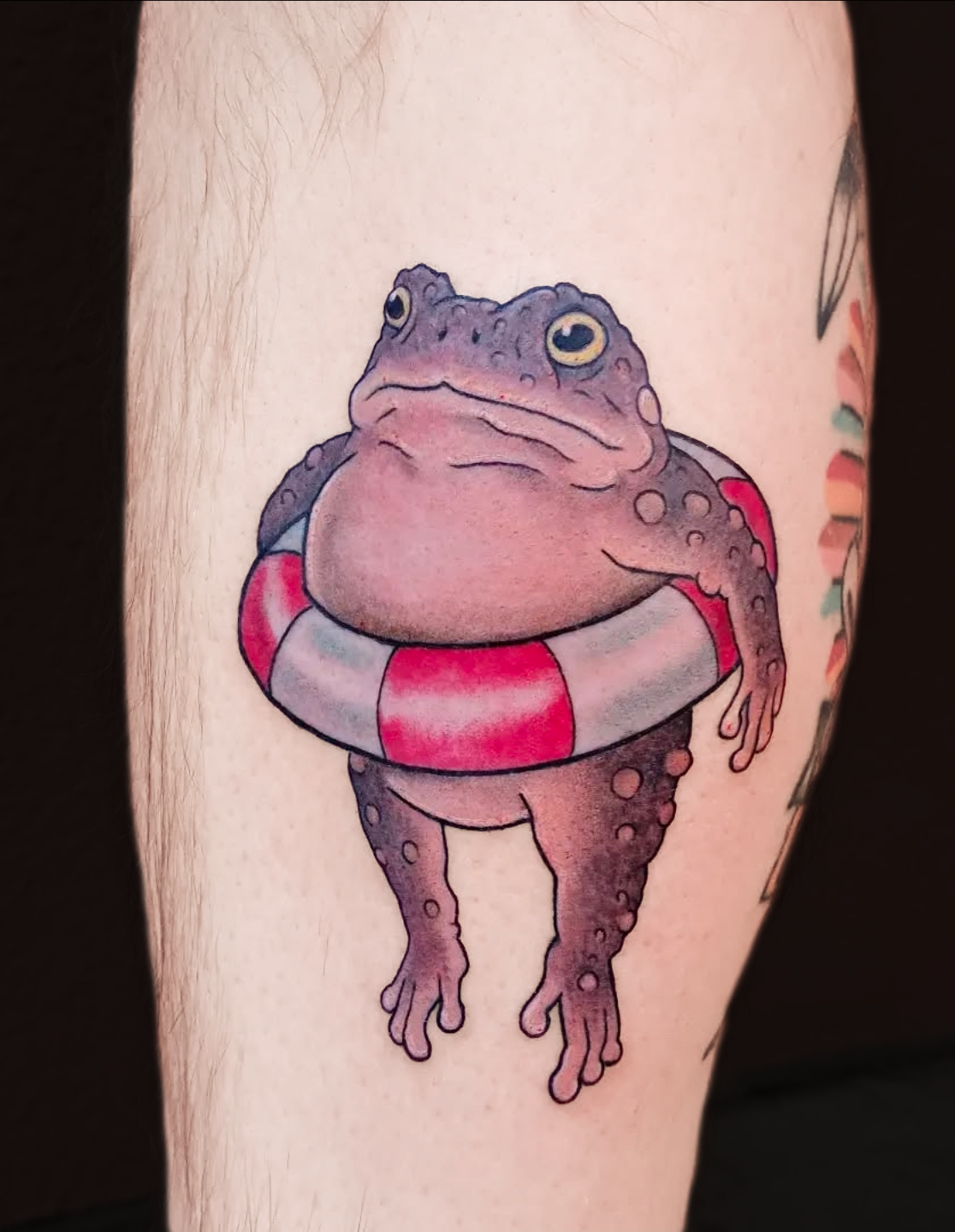 Tattoo of a frog wearing a red and white swim ring, with a relaxed expression, on someone's arm.