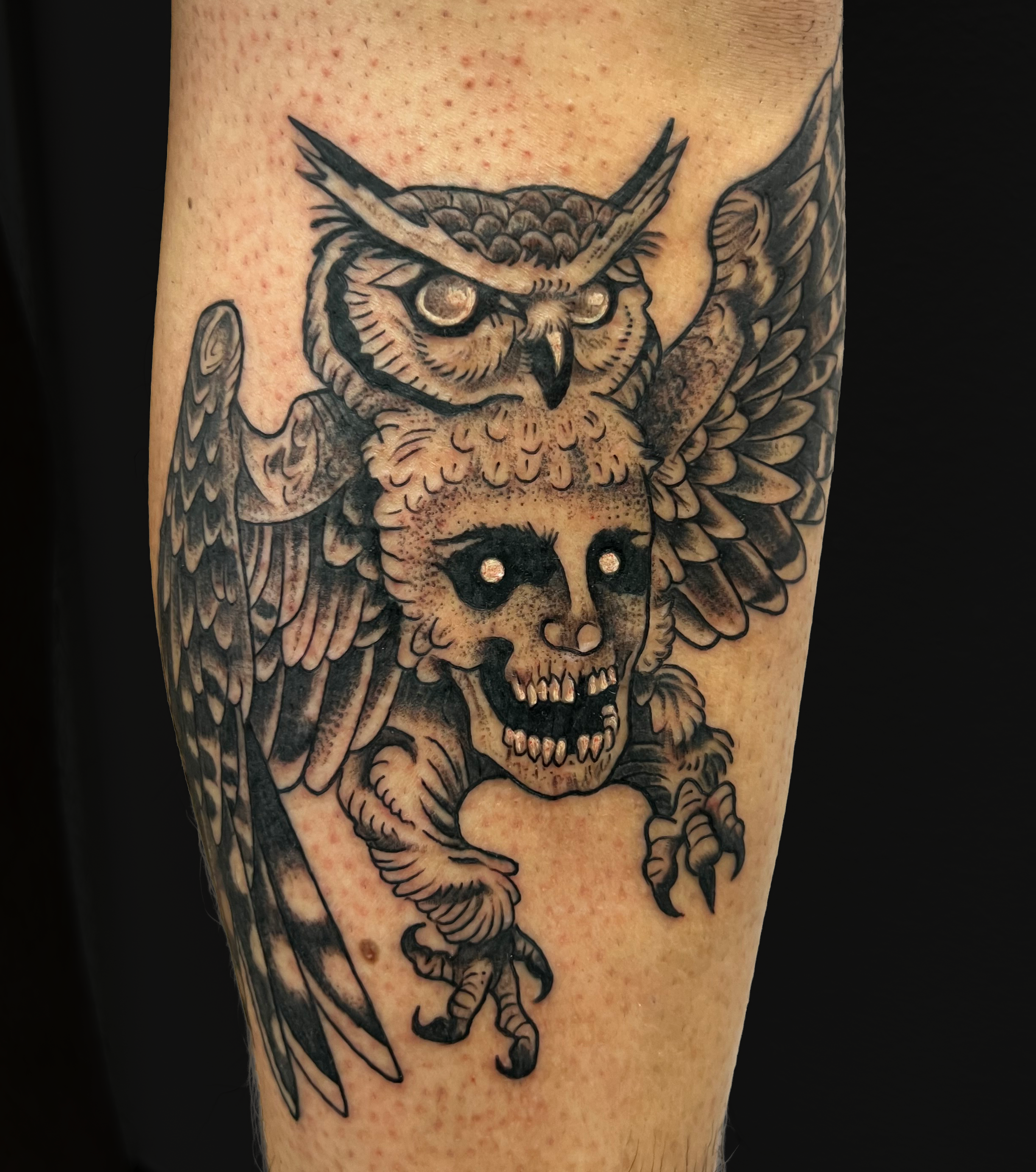 Black and gray tattoo of an owl with outstretched wings, clutching a skull in its claws, on person's arm.
