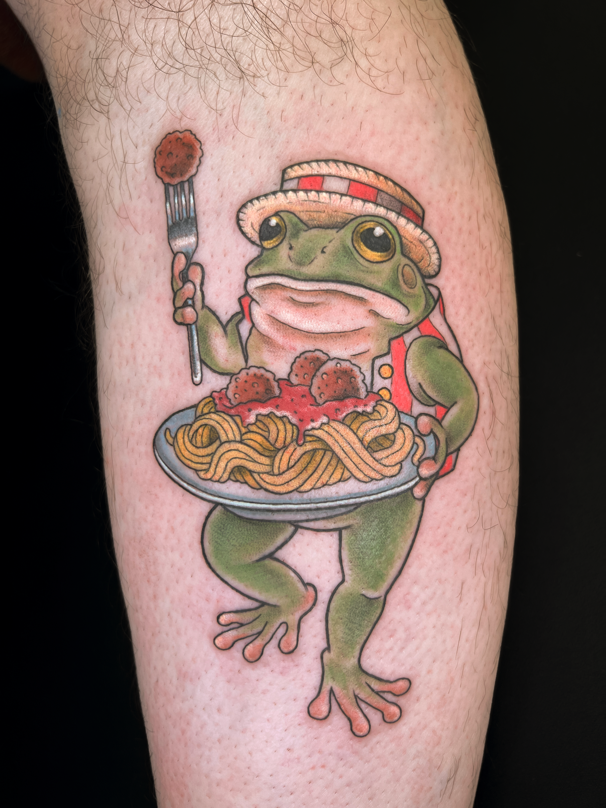Tattoo of a frog wearing a straw hat, holding a plate of spaghetti with meatballs, with a fork in one hand and a red vest, on a person's arm.