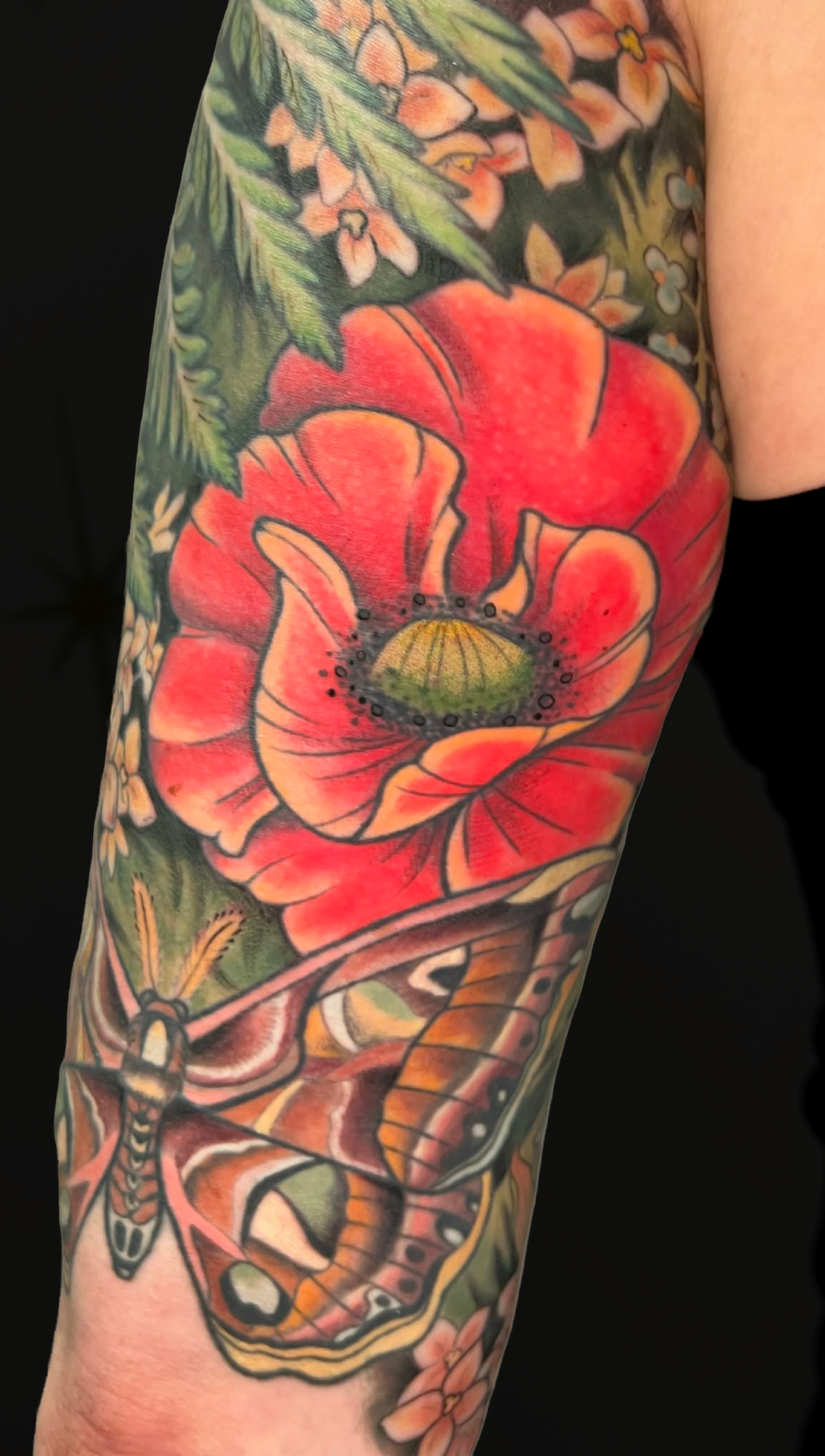 Tattoo of a large orange and yellow flower with green leaves on a person’s arm, accompanied by other floral and nature-themed designs.
