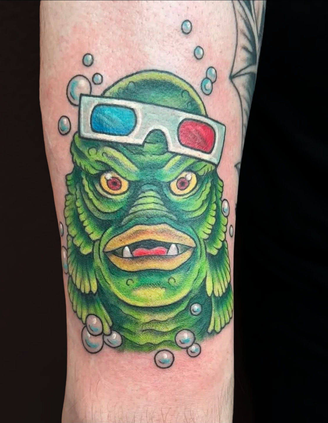 Tattoo of a mythological creature with green skin, yellow eyes, and fanged mouth, wearing 3D glasses, surrounded by bubbles.