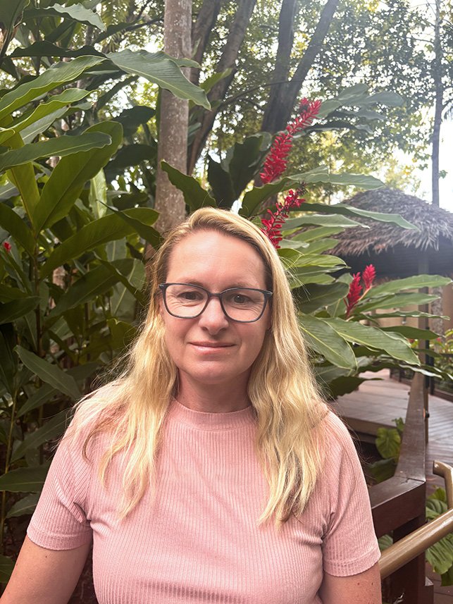 Woman with long blonde hair, glasses, wearing a pink shirt, standing outdoors with green foliage and red flowers behind her.