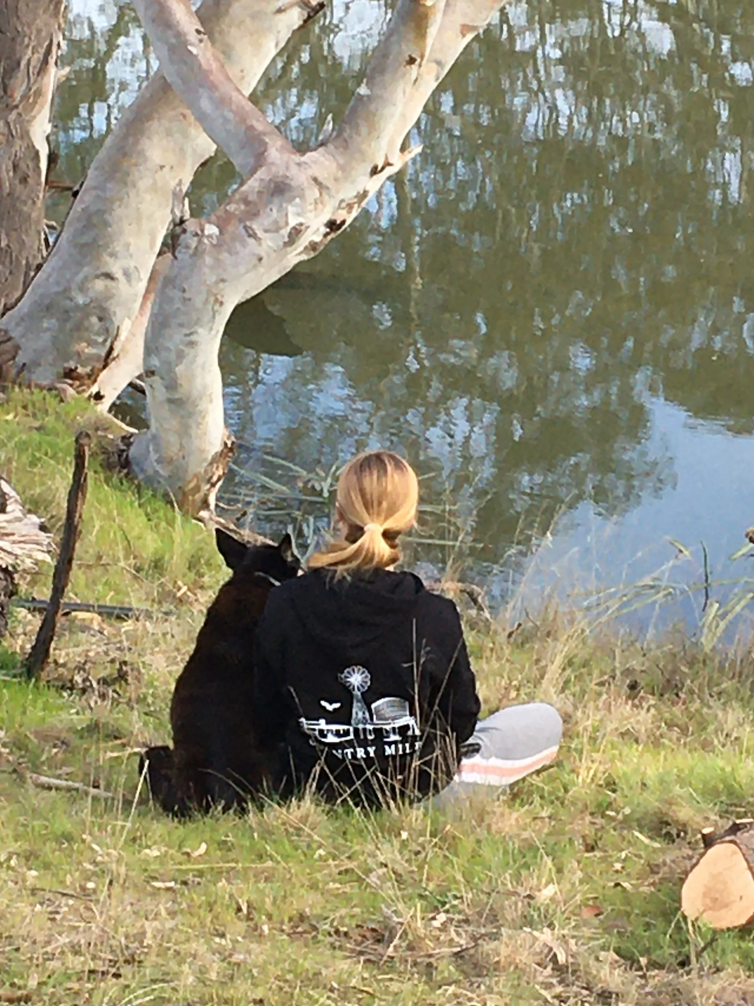 A person with blonde hair wearing a black hoodie with white graphics and white leggings with orange and pink stripes, sitting on grass near a body of water, with a black and white dog nearby, surrounded by trees.