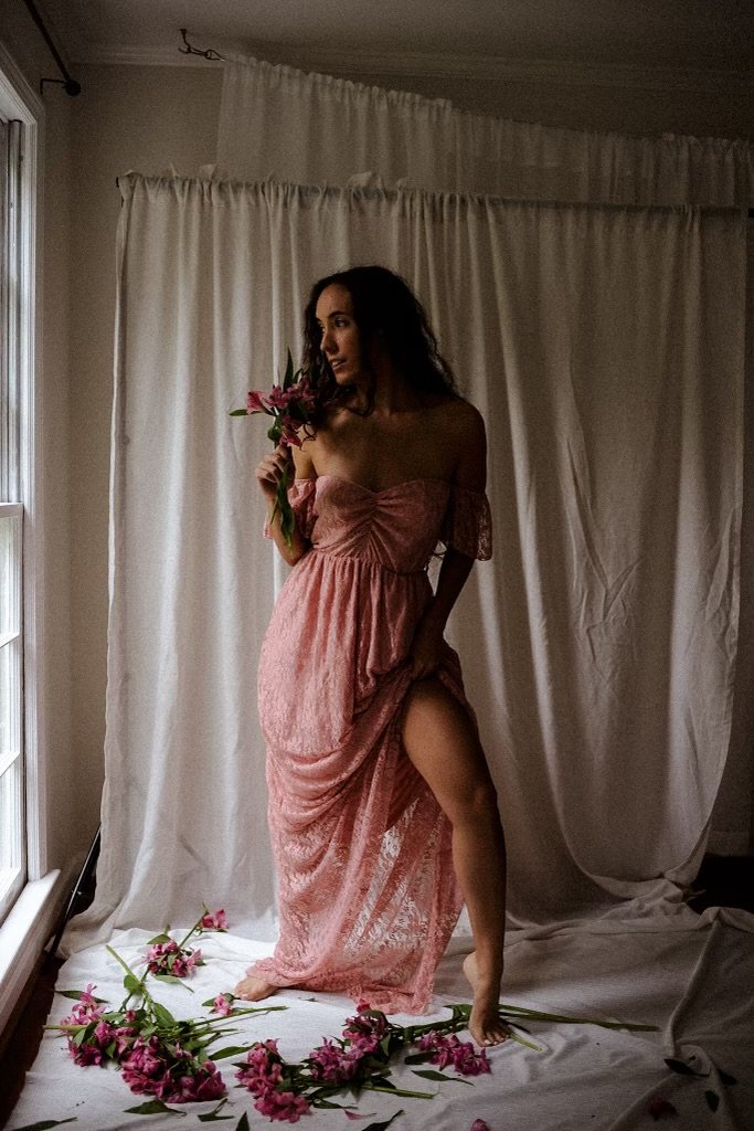 Woman in a pink lace dress holding flowers, standing in front of white curtains with flowers on the floor and in her hand.