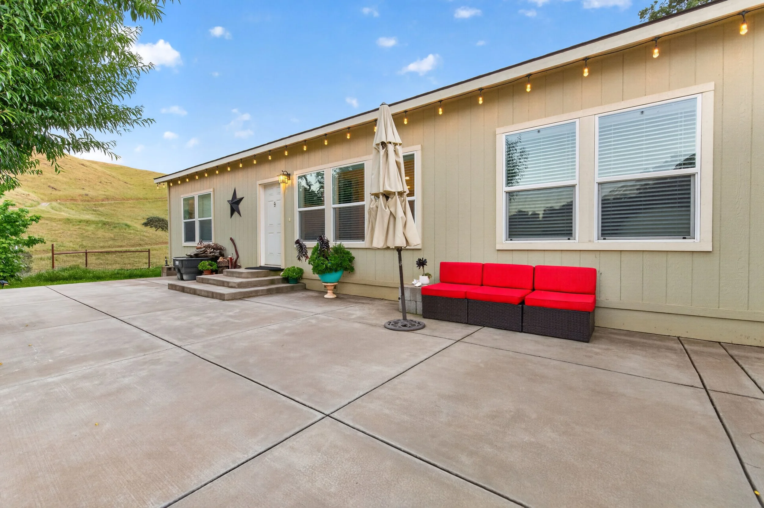 A backyard patio with concrete paving, a red outdoor sofa, a closed umbrella, potted plants, and a beige house with string lights and hillside in the background.