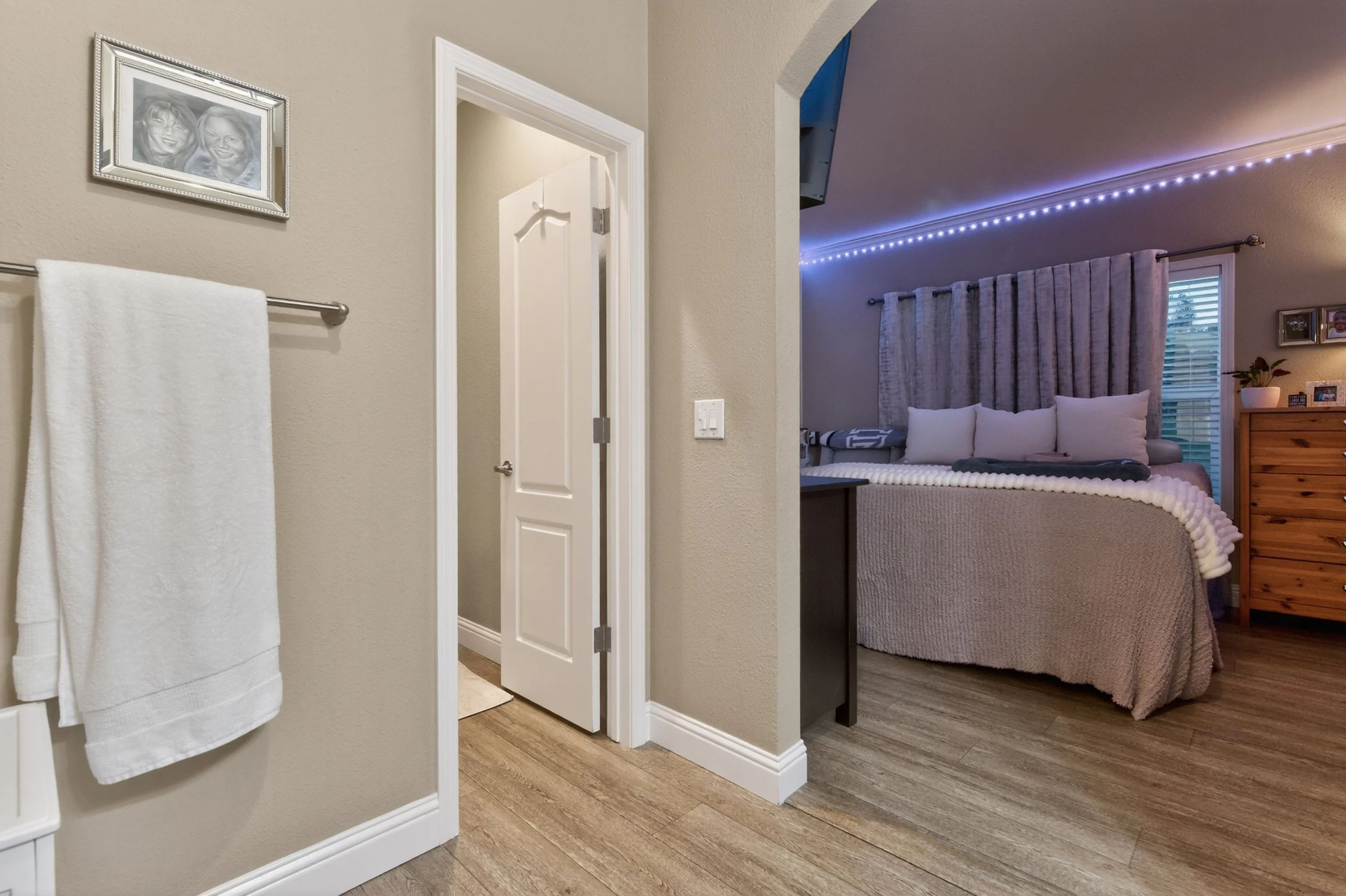 A bedroom with a bed, a window with curtains, a wooden dresser, and string lights on the ceiling. Part of a bathroom door and a towel on a towel rack are visible in the foreground.