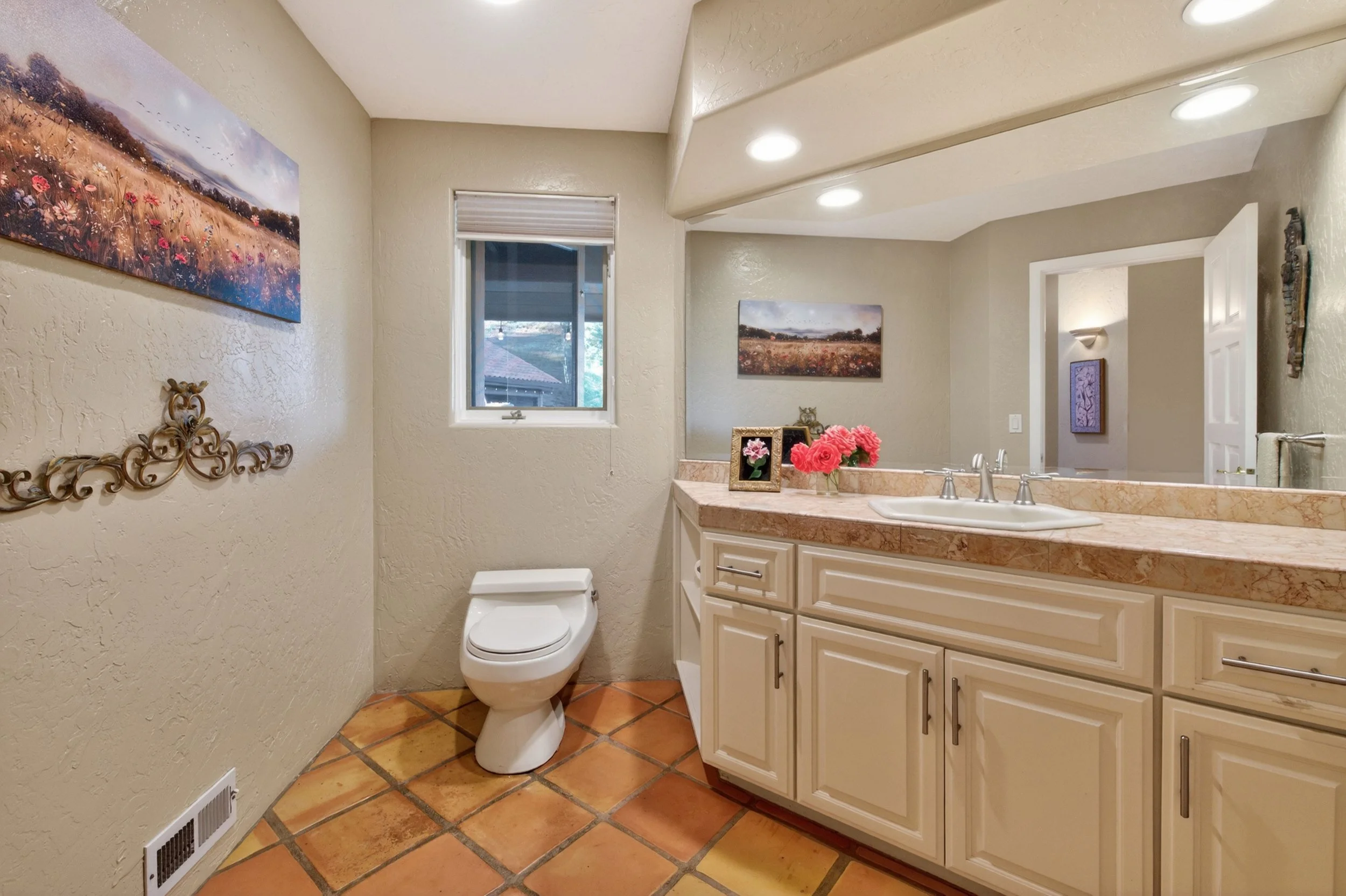 A small bathroom with a beige color scheme. There is a toilet on the left, above which is a painting of a field with wildflowers. To the right, there is a vanity with a marble countertop, a sink, flowers, and framed photos. Above the vanity, there is