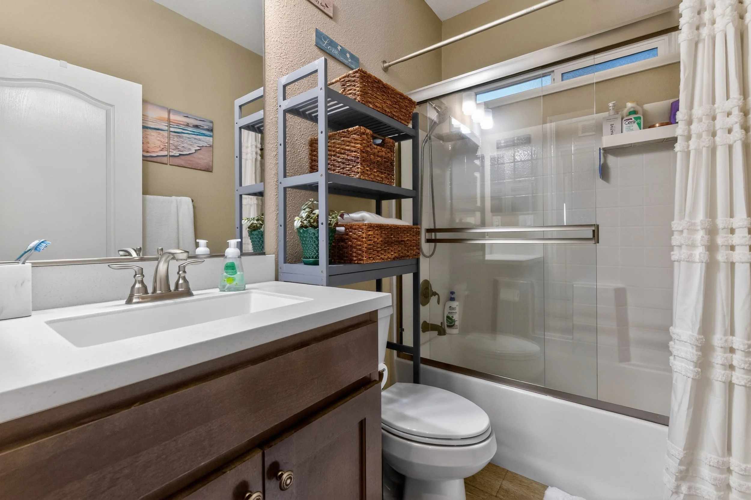 A bathroom with a vanity, a mirror, a toilet, and a glass-enclosed shower with a white curtain, wooden flooring, and wall art.