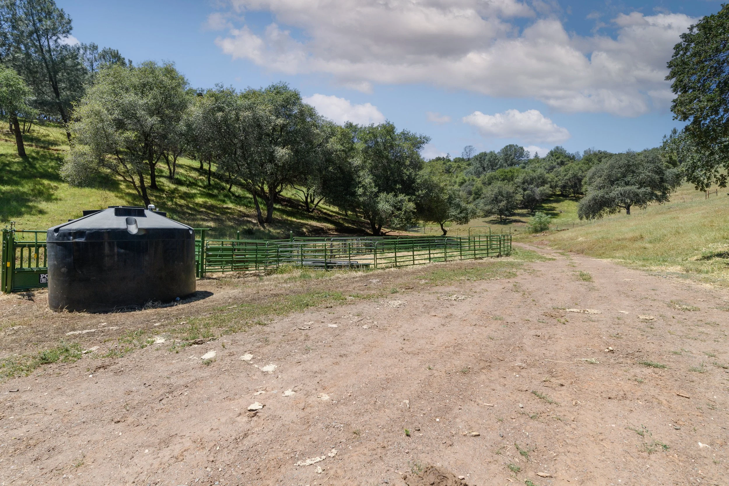 A farm or ranch landscape with green trees on a hillside, a dirt path, and a water tank in the foreground. The sky is partly cloudy with patches of blue.