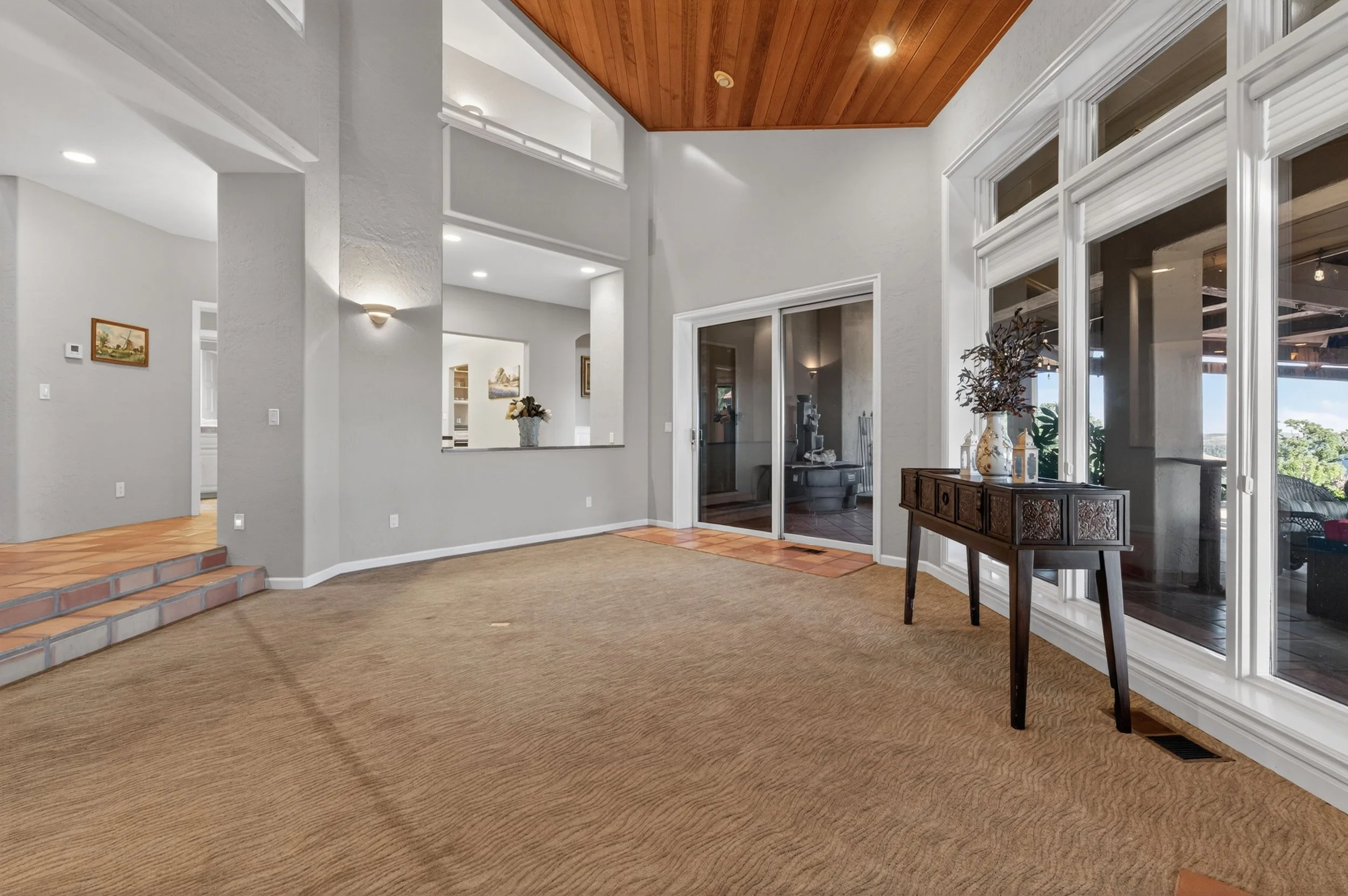 Living room with beige carpet, white walls, large windows, and wooden ceiling. A small wooden table with decorative items and a vase with dried flowers are near the window, with a sliding glass door leading outside.