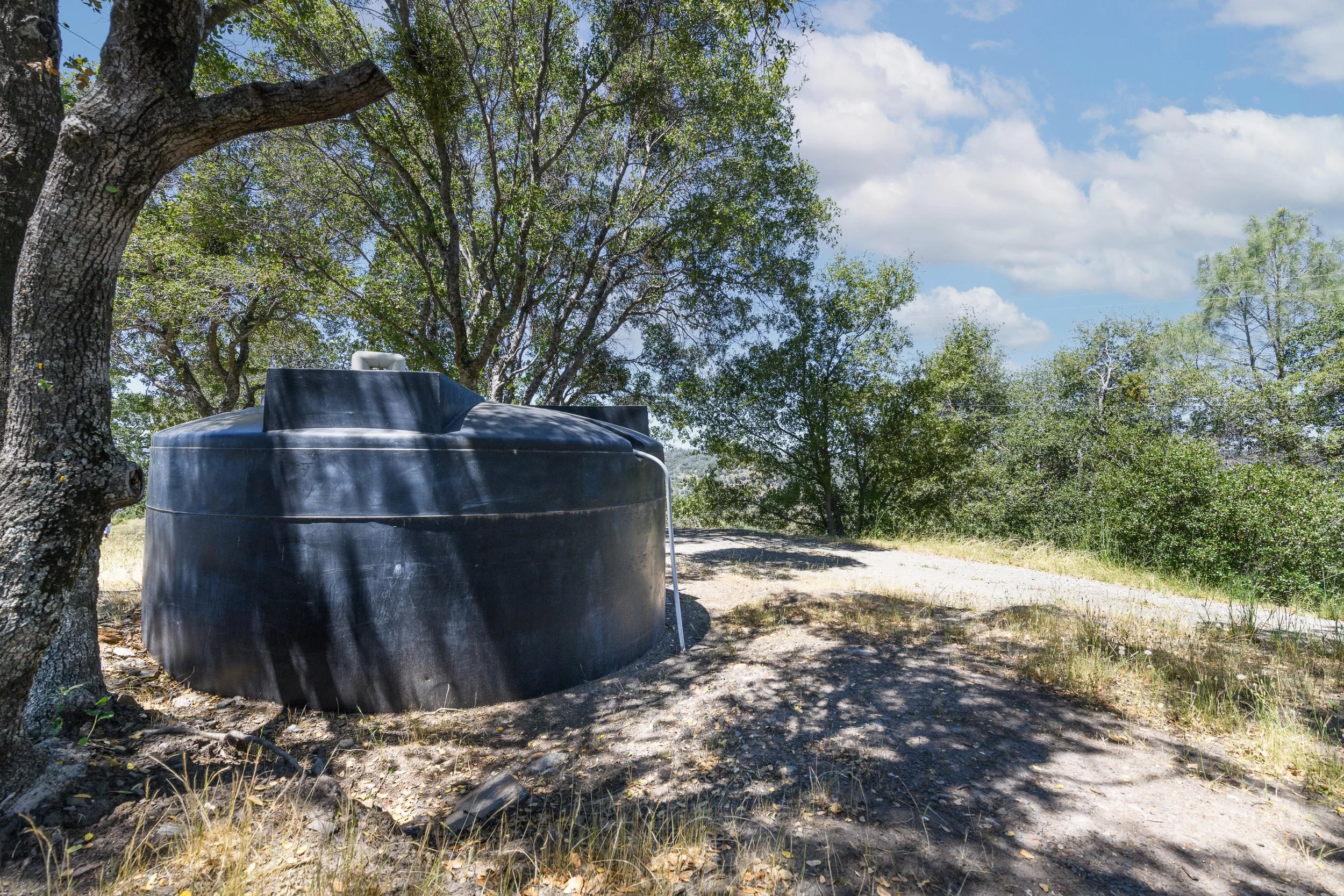 A large black water storage tank outdoors in a natural setting surrounded by trees, with a partly cloudy sky overhead.