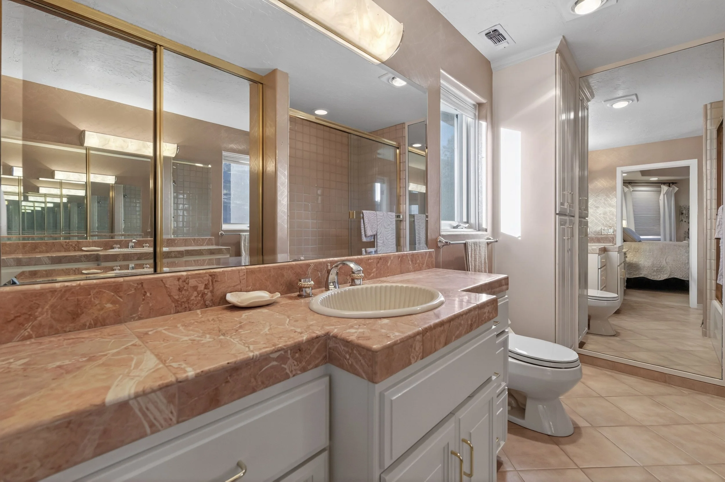 A bathroom with marble countertop, a sink, and a large mirror. There are towels hanging on a towel rack, a window letting in natural light, a toilet, a shower with glass doors, and a separate room with a bed visible through the doorway.