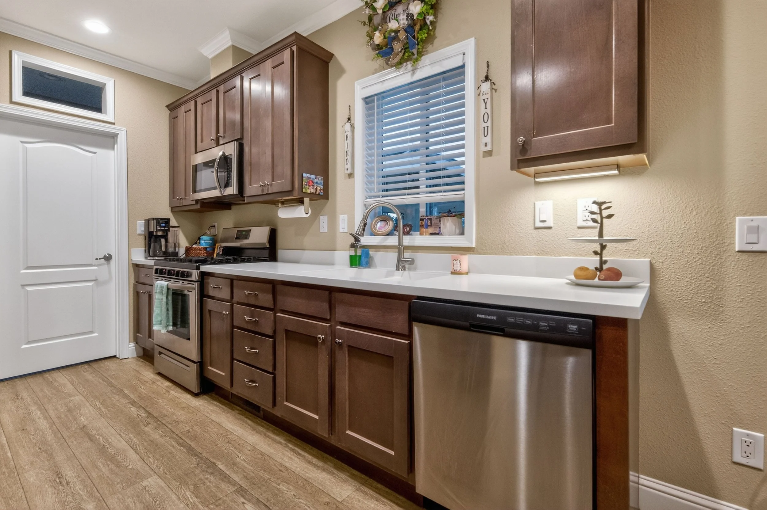 Kitchen with brown cabinets, stainless steel dishwasher and microwave, white counter, window with blinds, and decorative wreath and signs on wall.